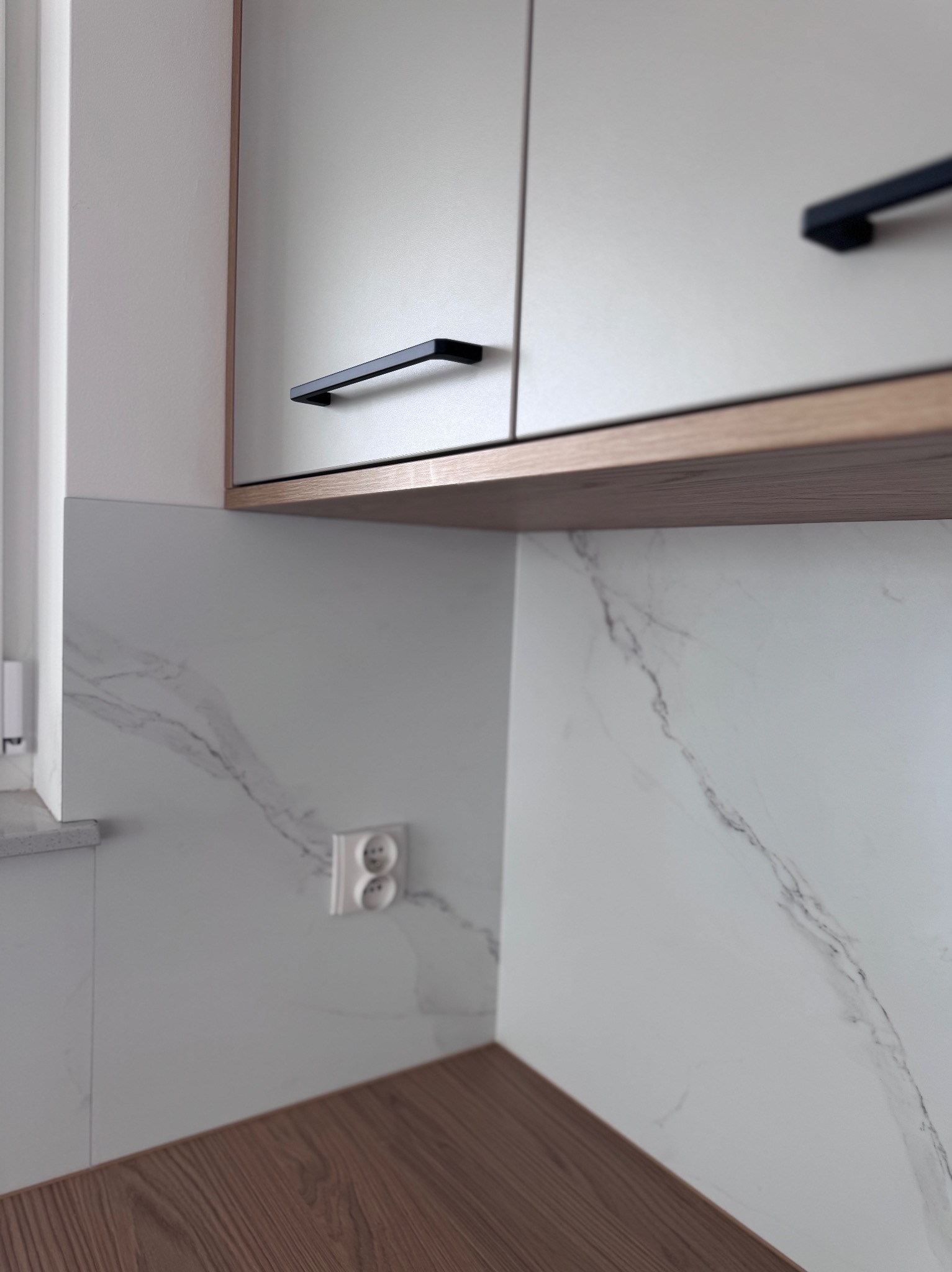 Fragment nowoczesnej kuchni: białe szafki z czarnymi uchwytami, drewniany blat i marmurowe panele ścienne z gniazdkiem elektrycznym. Minimalistyczny design.