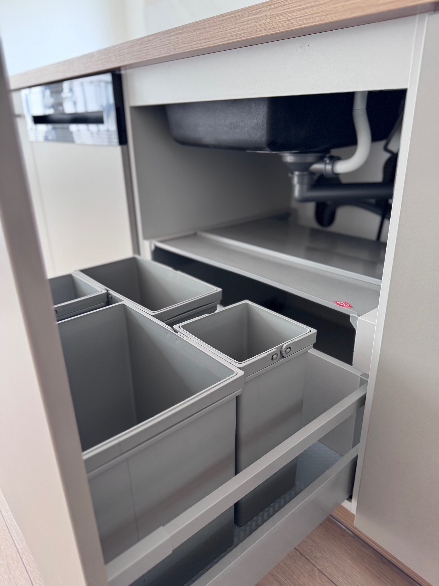 Szafka kuchenna z wysuwanym systemem sortowania odpadów, widoczny zlew i instalacja. Jasne kolory, minimalistyczny design.