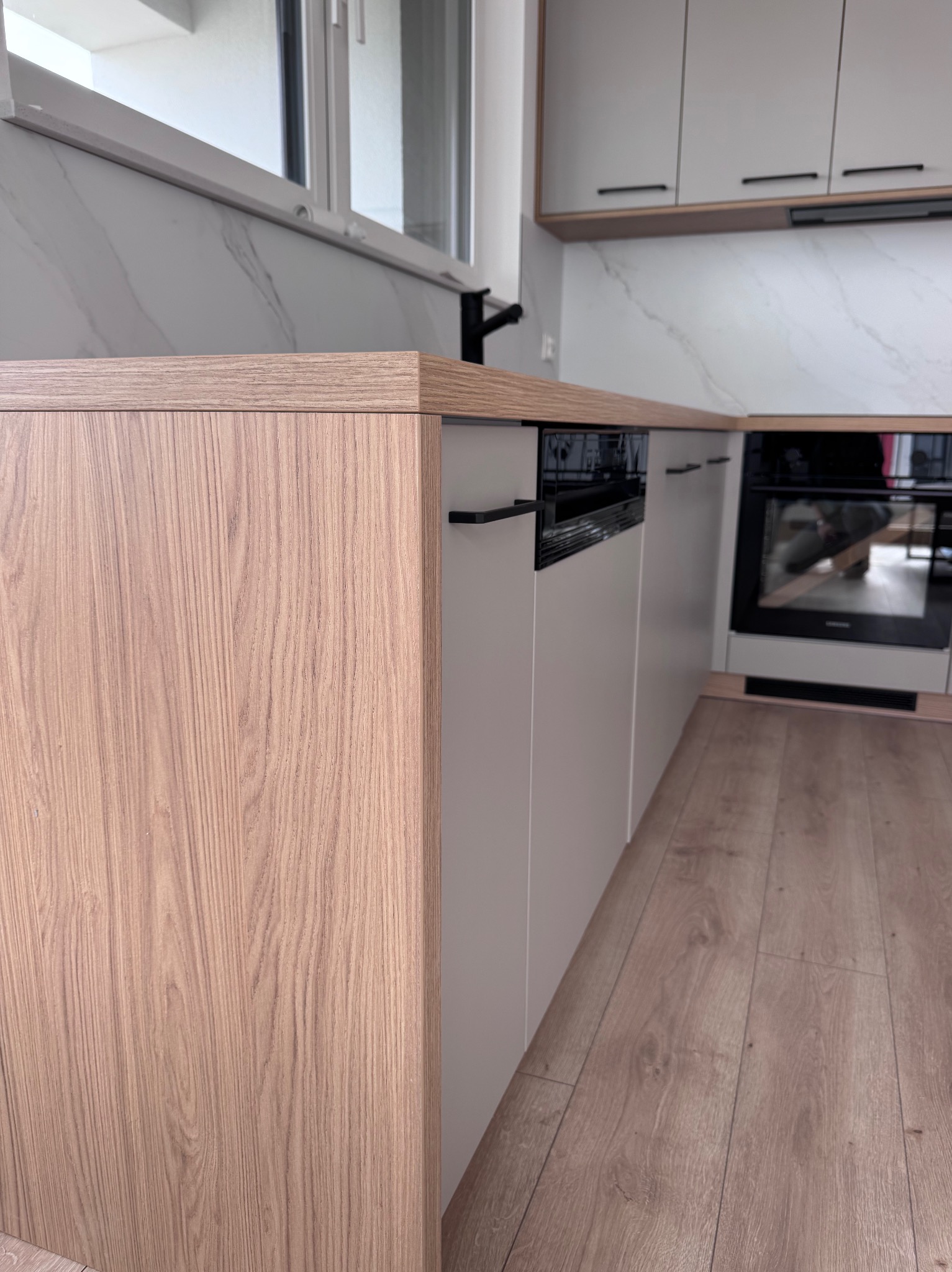 Nowoczesna kuchnia z drewnianymi frontami i blatem, szare szafki, marmurowy backsplash i drewniana podłoga. Widoczny piekarnik i minimalistyczne uchwyty.