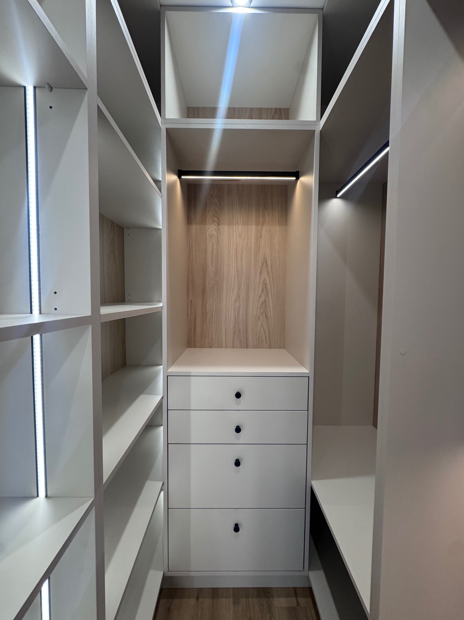 Wnętrze szafy wnękowej z oświetleniem LED, jasne półki i szuflady z ciemnymi uchwytami, drewniane wykończenie w środkowej części. Funkcjonalne rozwiązanie do przechowywania.