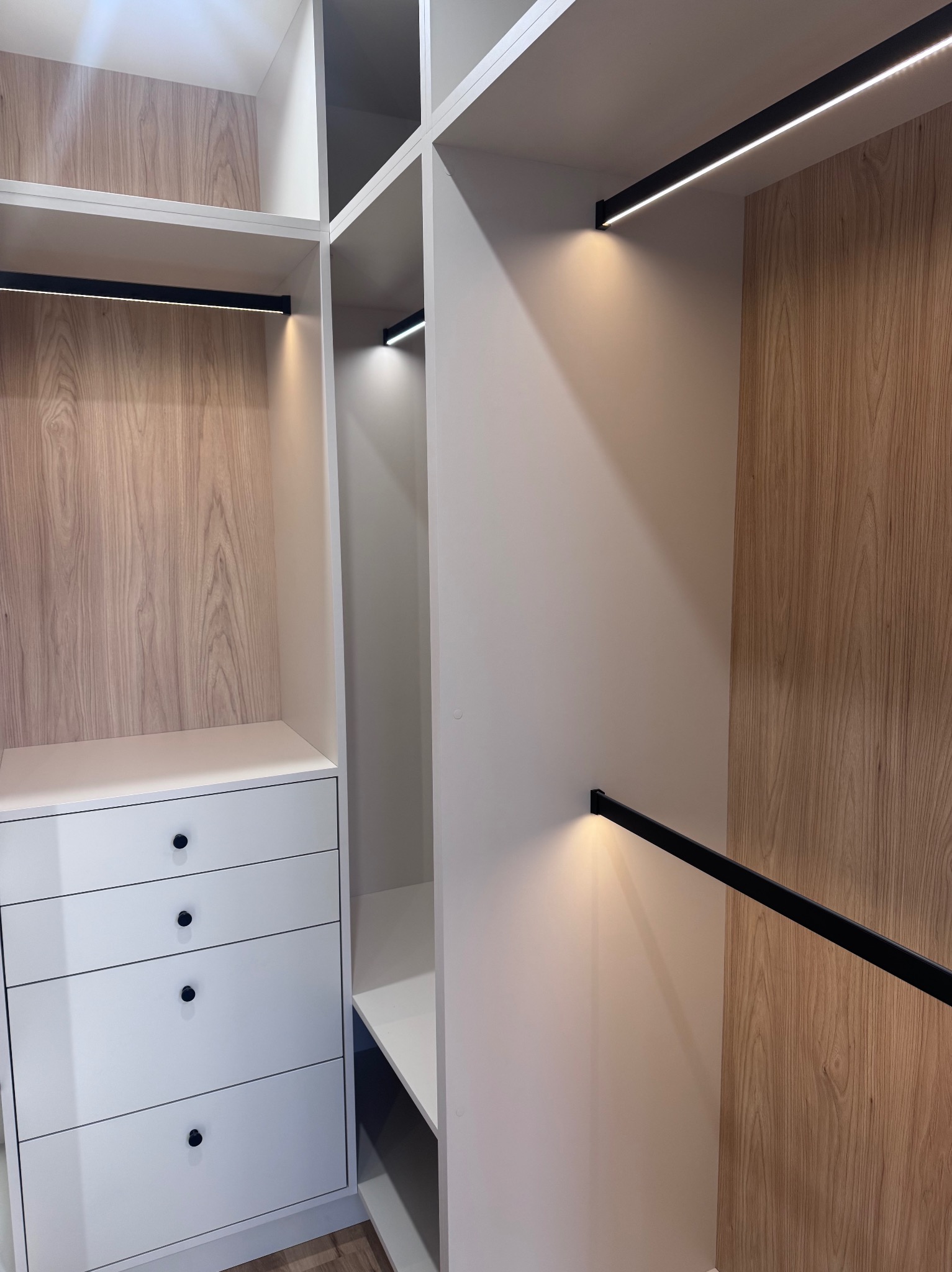 Wnętrze szafy wnękowej z oświetleniem LED, białe fronty szuflad z czarnymi gałkami, drewnopodobne panele na ścianach, minimalistyczny design.