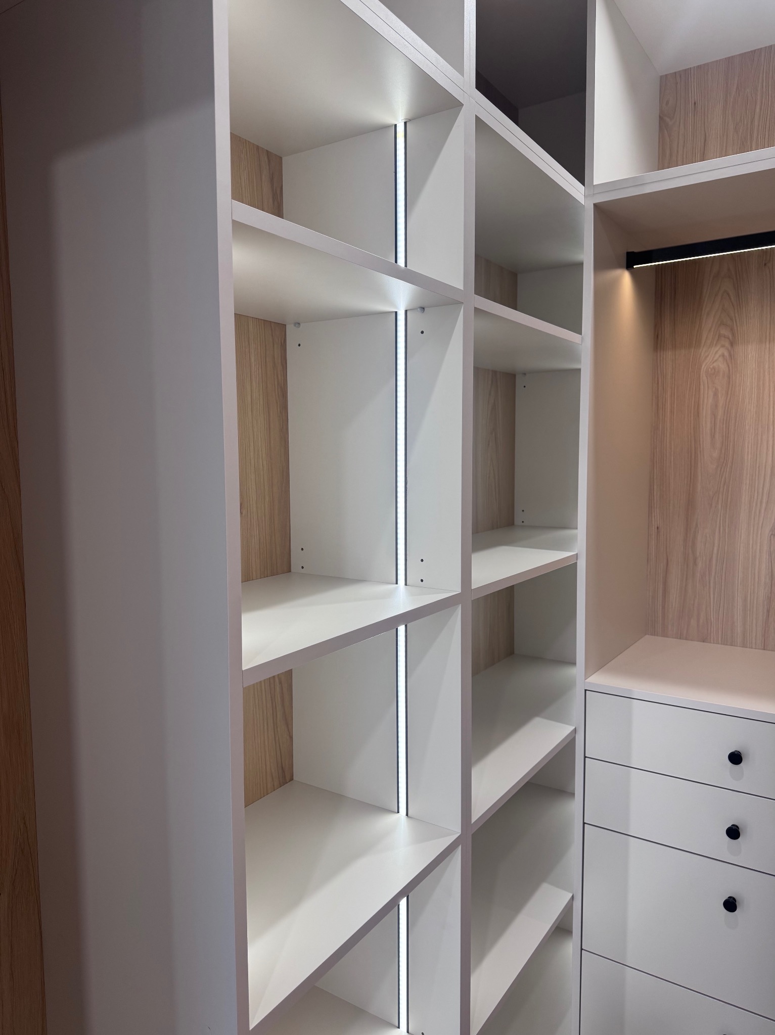 Biała szafa wnękowa z oświetleniem LED i drewnianym wykończeniem, widok z bliska na półki i szuflady z czarnymi gałkami. Nowoczesny design.