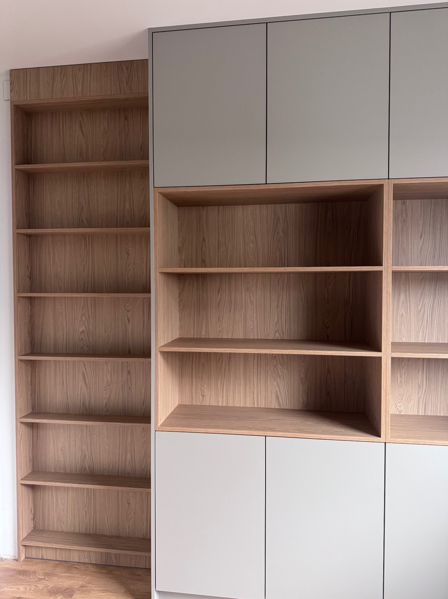 Wysoka zabudowa biurowa z szarymi szafkami i regałami z jasnego drewna. Widoczne półki i fronty szafek. Minimalistyczny design, nowoczesne wykończenie.