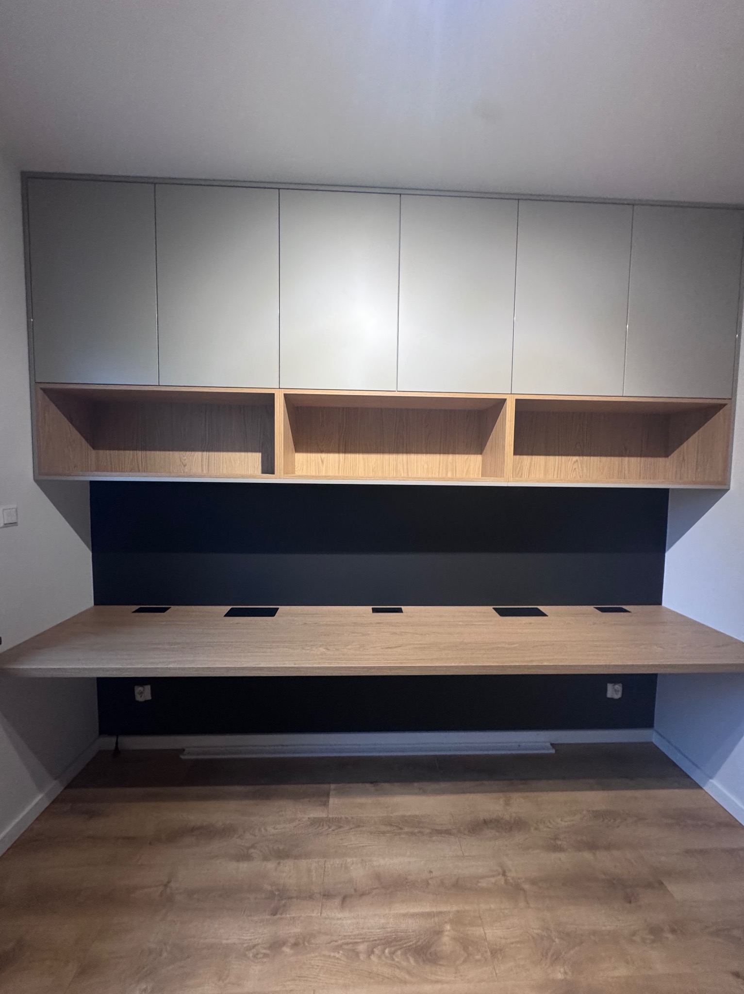 Nowoczesne biurko na ścianie z szafkami wiszącymi w odcieniach szarości i drewna, z czarnym panelem na ścianie i otworami na kable, na tle podłogi z paneli.