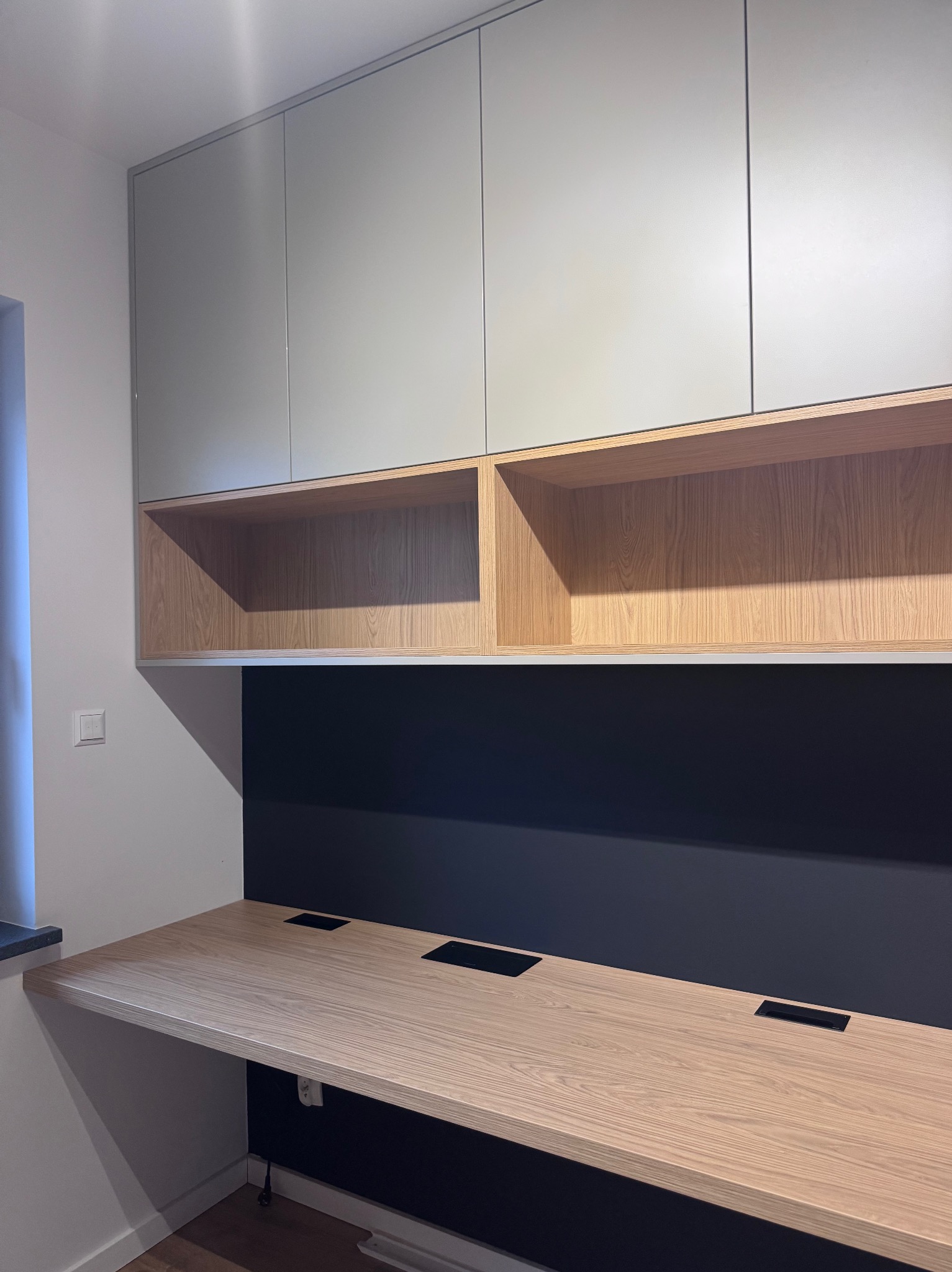 Nowoczesne biurko z drewnianym blatem i szarymi szafkami wiszącymi. Blat z wbudowanymi gniazdami. Czarna ściana za biurkiem. Minimalistyczny design.