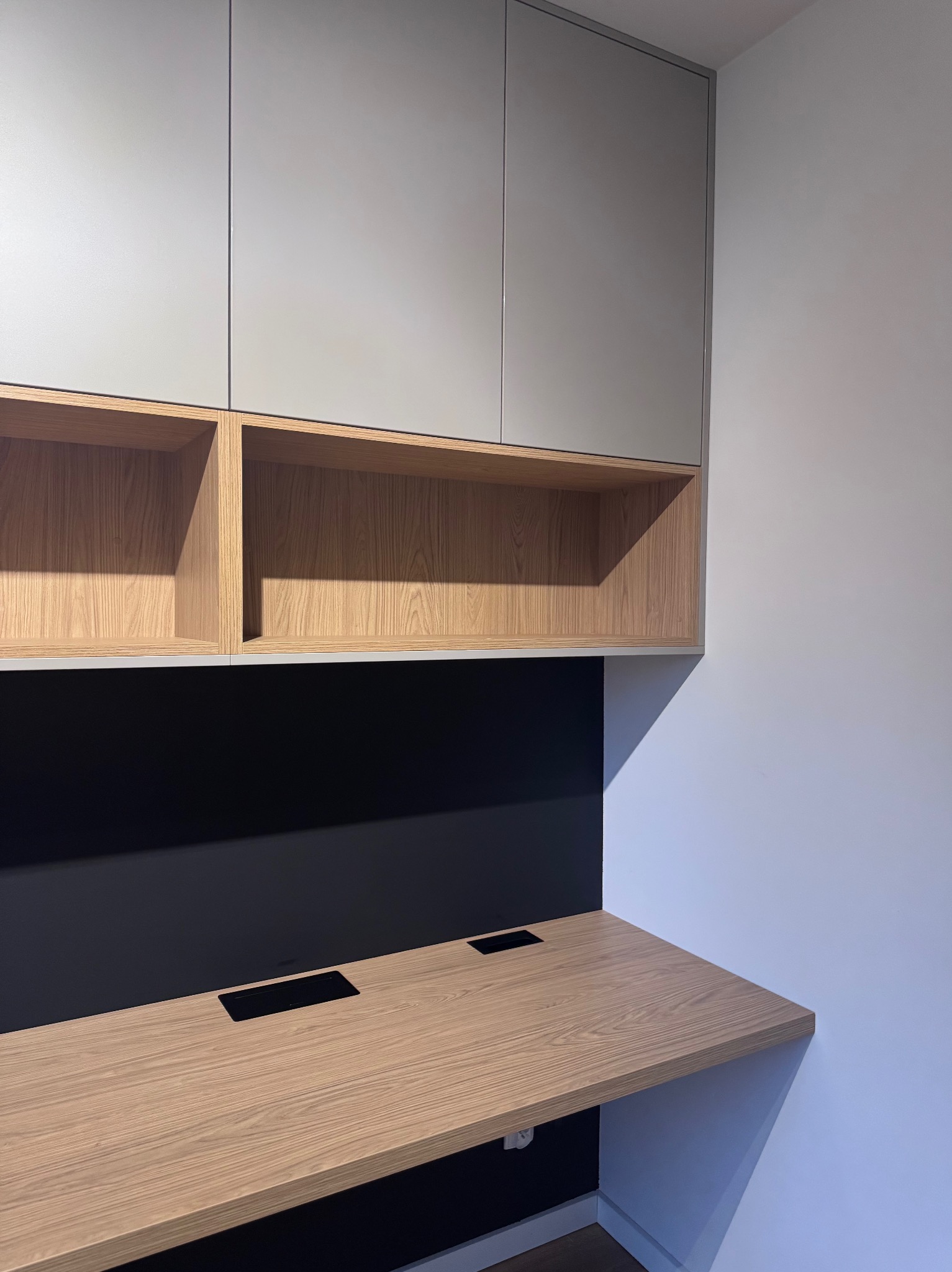 Nowoczesne biurko z drewnianym blatem i szafkami wiszącymi w odcieniach szarości i drewna, z czarnym panelem ściennym. Minimalistyczny design, gniazda w blacie.