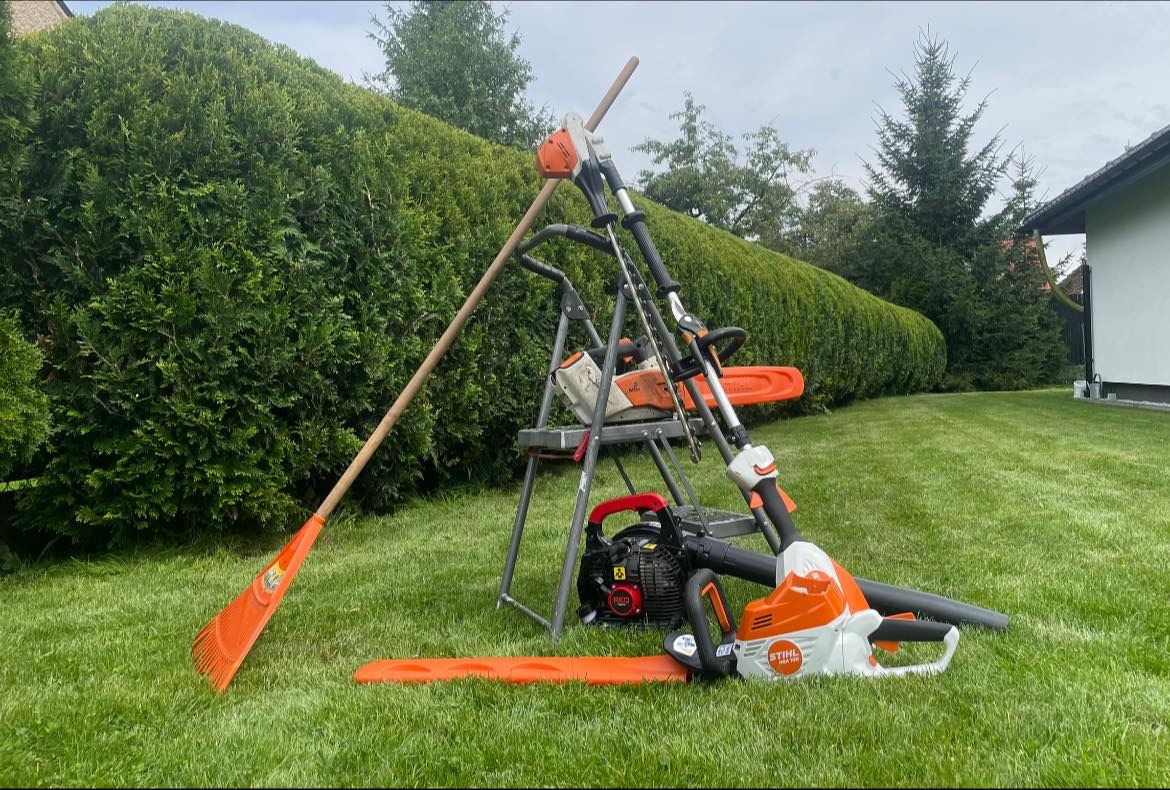 Zestaw narzędzi ogrodniczych Stihl: dmuchawa, nożyce do żywopłotu, pilarka na drabinie, grabie wachlarzowe na trawniku przed żywopłotem.