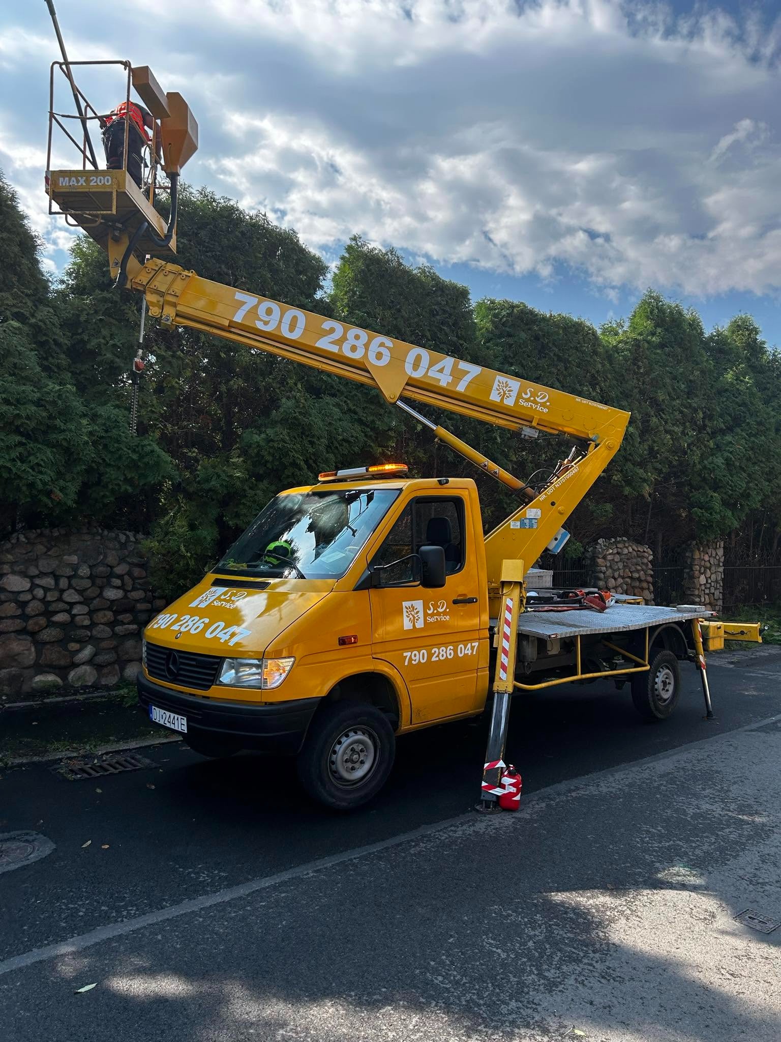 Żółty podnośnik koszowy z numerem telefonu na tle zielonych drzew i pochmurnego nieba. W koszu widoczna osoba w pomarańczowym kombinezonie.