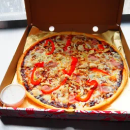 Pizza z kurczakiem, szynką i papryką w kartonie na stole, obok sos w pojemniku. Widok z góry, jasne światło.