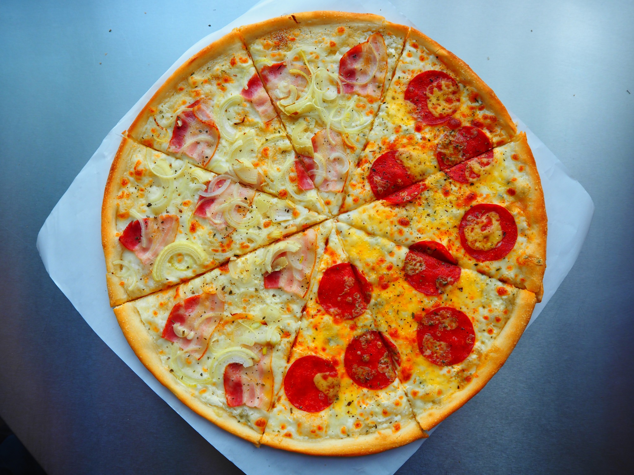 Pizza 'pół na pół' z pepperoni i szynką z cebulą, podzielona na trójkątne kawałki, leżąca na papierze do pieczenia. Widok z góry, ostre światło.