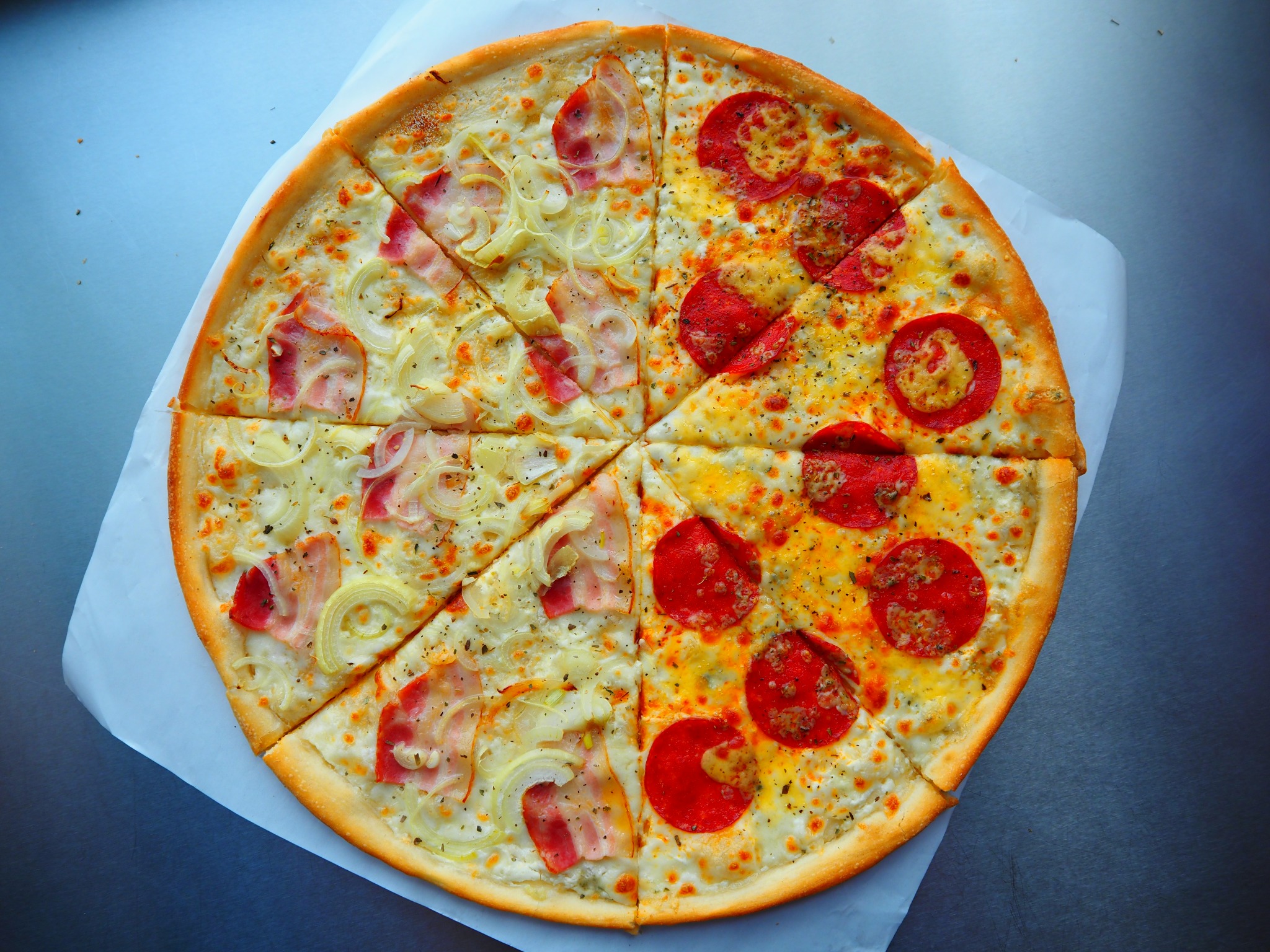 Pizza na białym papierze z dwoma rodzajami dodatków: salami i bekon z cebulą, podzielona na równe kawałki. Widok z góry, ostre światło.