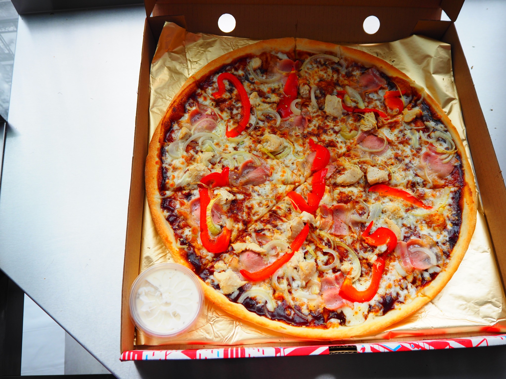 Pizza z kurczakiem, szynką, czerwoną papryką i cebulą w kartonie na złotym papierze, z sosem obok. Widok z góry.