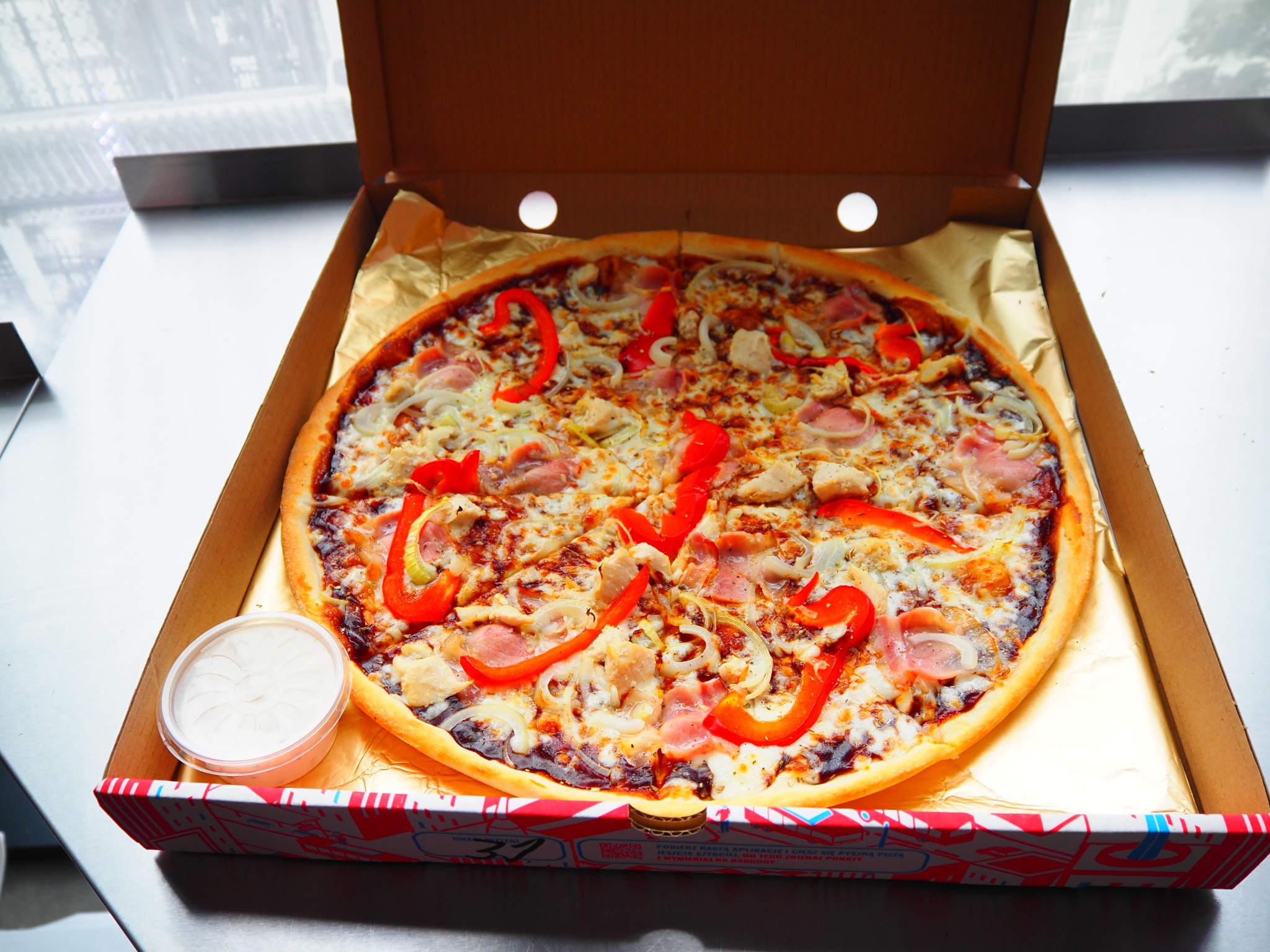 Pizza z kurczakiem, szynką i papryką w kartonie na stole, obok sos w pojemniku. Widok z góry, jasne światło.