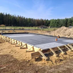 Przygotowany fundament z izolacją, szalunkiem i częściowo widocznym zbrojeniem na placu budowy w słoneczny dzień. W tle drzewa i wykopana ziemia.