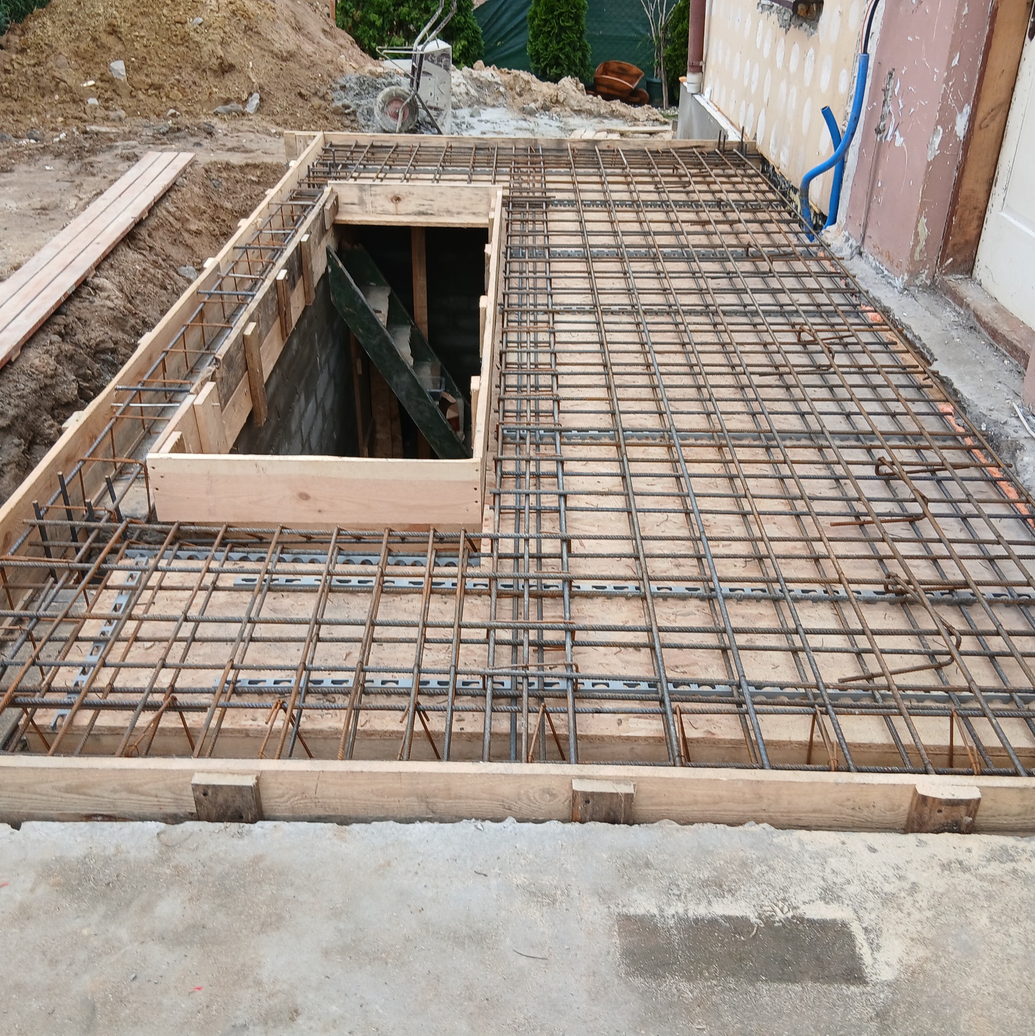 Zbrojenie płyty fundamentowej z otworem, przygotowane do zalania betonem. Widoczne drewniane szalunki i stalowe pręty tworzące siatkę zbrojeniową.