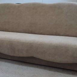 Beżowa sofa z zaokrąglonymi podłokietnikami i falistym oparciem, ustawiona na betonowej podłodze w jasnym pomieszczeniu. Widoczny fragment stołu warsztatowego.