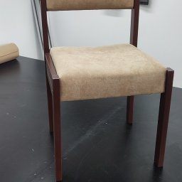 Krzesło z beżową tapicerką i ciemnobrązową ramą na tle próbek tkanin. Widoczne detale wykończenia i materiały użyte do renowacji krzesła.