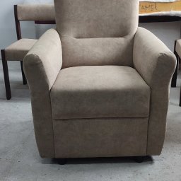 Beżowy, tapicerowany fotel z wysokim oparciem na tle warsztatu tapicerskiego. W tle widoczne inne meble w trakcie renowacji.