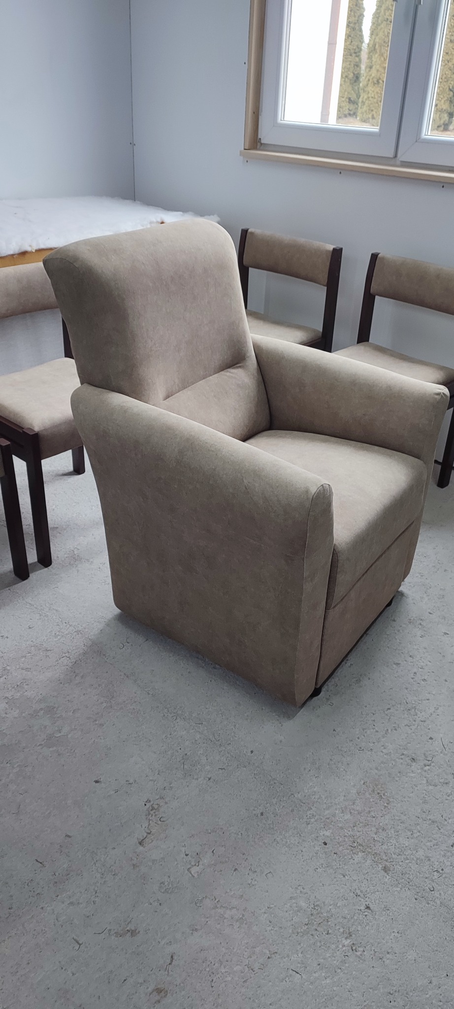 Beżowy fotel z podłokietnikami na tle krzeseł i okna. Fotel tapicerowany, o prostej formie, ustawiony na szarym tle. Widoczne detale wykończenia.