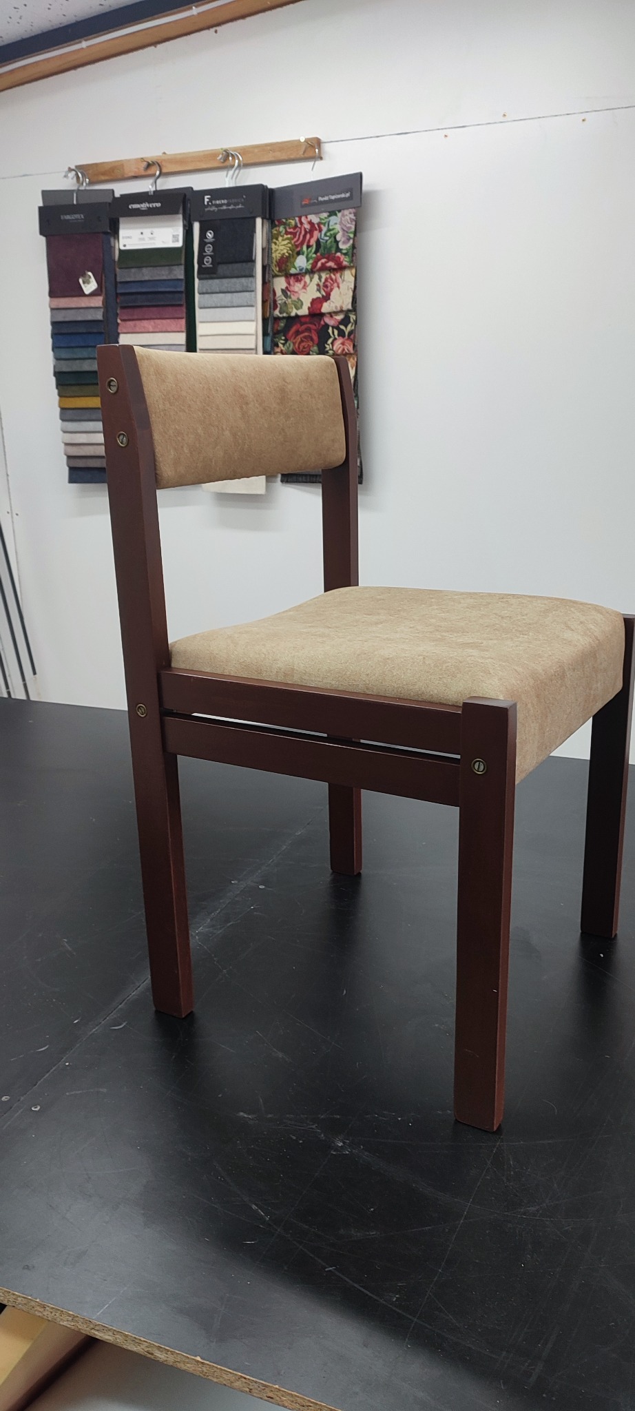 Krzesło tapicerowane beżową tkaniną na ciemnym blacie, w tle wzorniki materiałów. Widoczne detale wykończenia i odcienie brązu drewna.