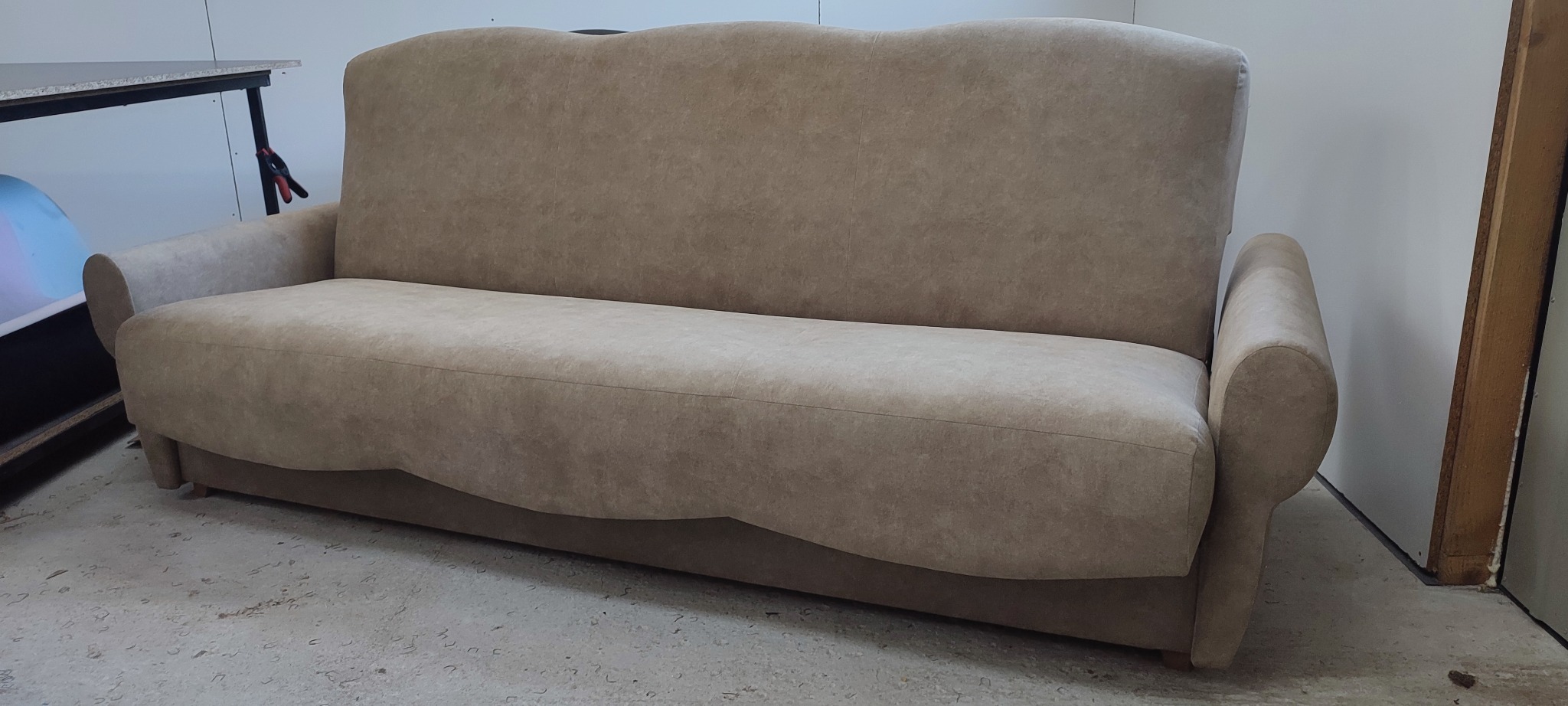Beżowa sofa z zaokrąglonymi podłokietnikami i falistym oparciem, ustawiona na betonowej podłodze w jasnym pomieszczeniu. Widoczny fragment stołu warsztatowego.
