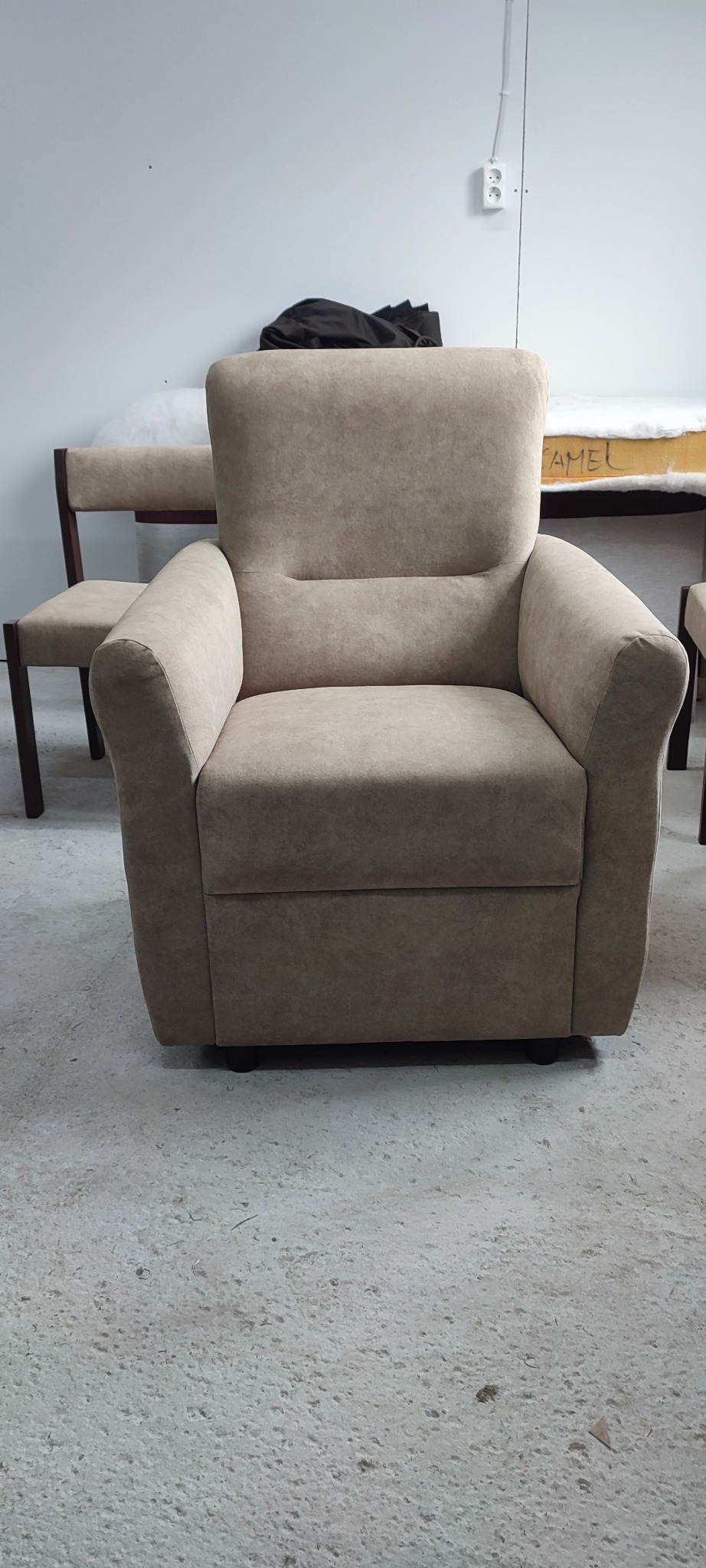 Beżowy, tapicerowany fotel z wysokim oparciem na tle warsztatu tapicerskiego. W tle widoczne inne meble w trakcie renowacji.