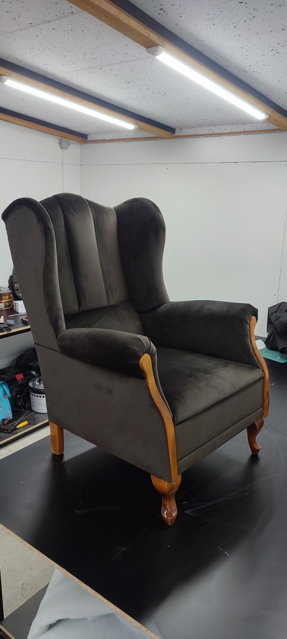 Ciemnobrązowy fotel typu uszak z drewnianymi elementami, po renowacji, stoi na czarnym blacie w warsztacie tapicerskim. Widoczne oświetlenie sufitowe.