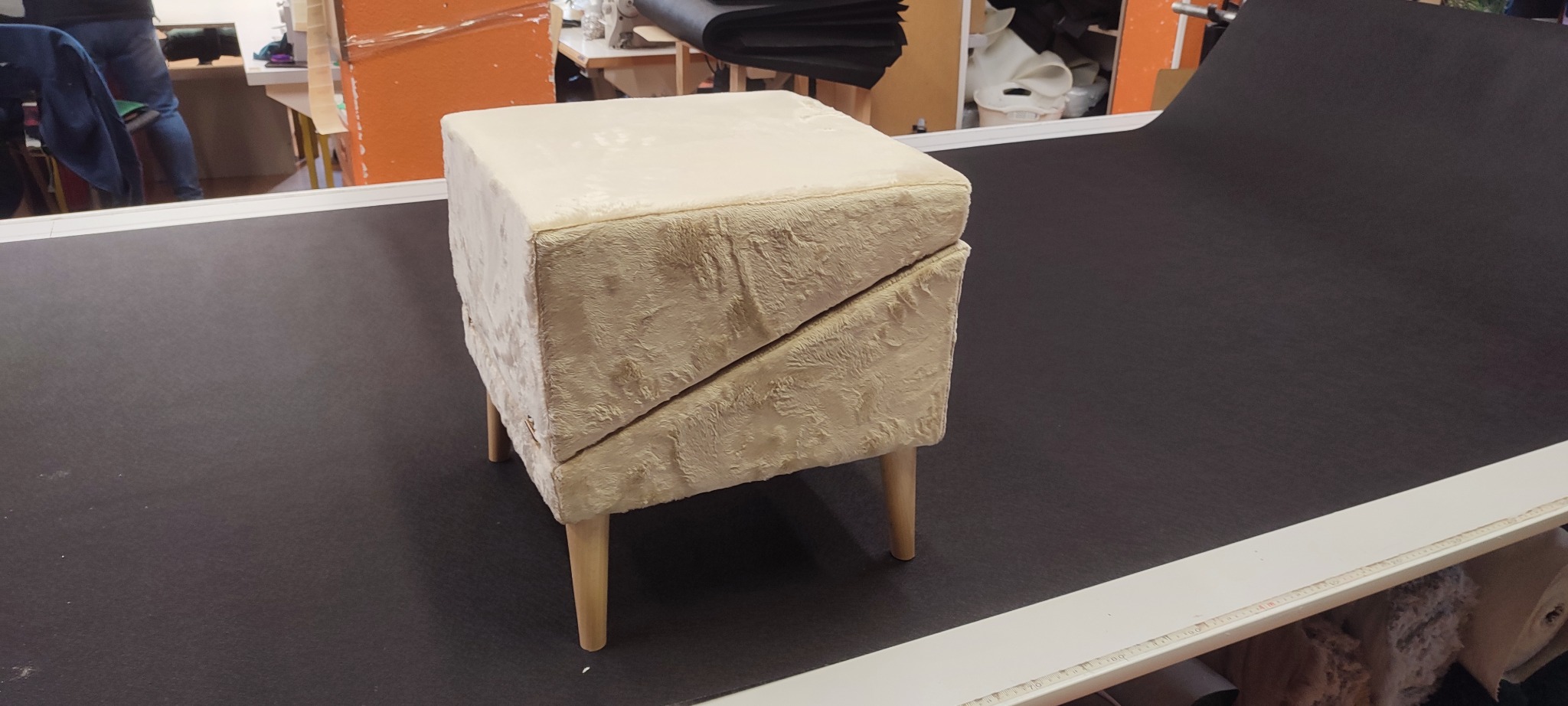 Beżowy, tapicerowany puf z drewnianymi nóżkami, ustawiony na czarnym materiale w warsztacie. Widoczne narzędzia i materiały w tle. Ujęcie z bliska, oświetlenie studyjne.