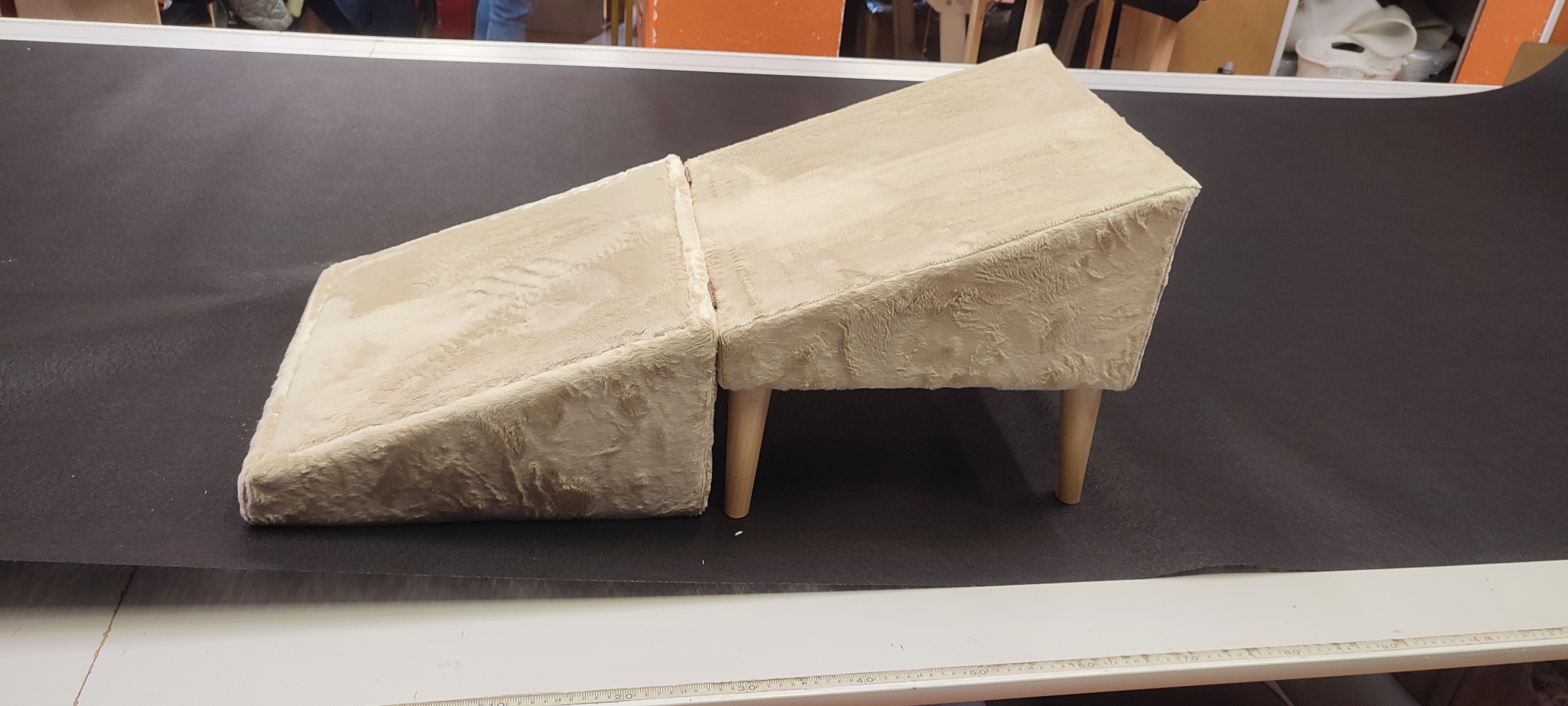 Beżowy, tapicerowany podnóżek z drewnianymi nóżkami, ustawiony na czarnym materiale. Widoczne szwy i faktura materiału. Ustawiony na stole z miarką.