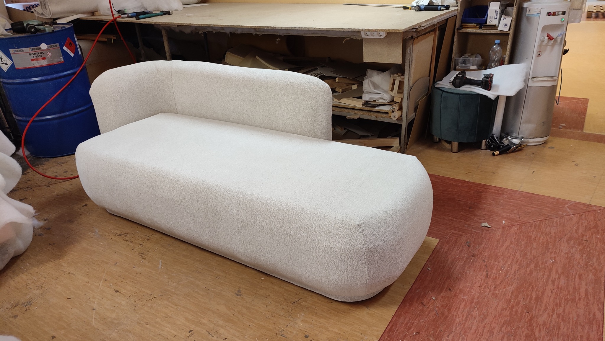 Nowoczesna, kremowa sofa tapicerowana bouclé w warsztacie, z widocznym niebieskim beczką i narzędziami w tle. Ujęcie z niskiego kąta podkreśla kształt mebla.