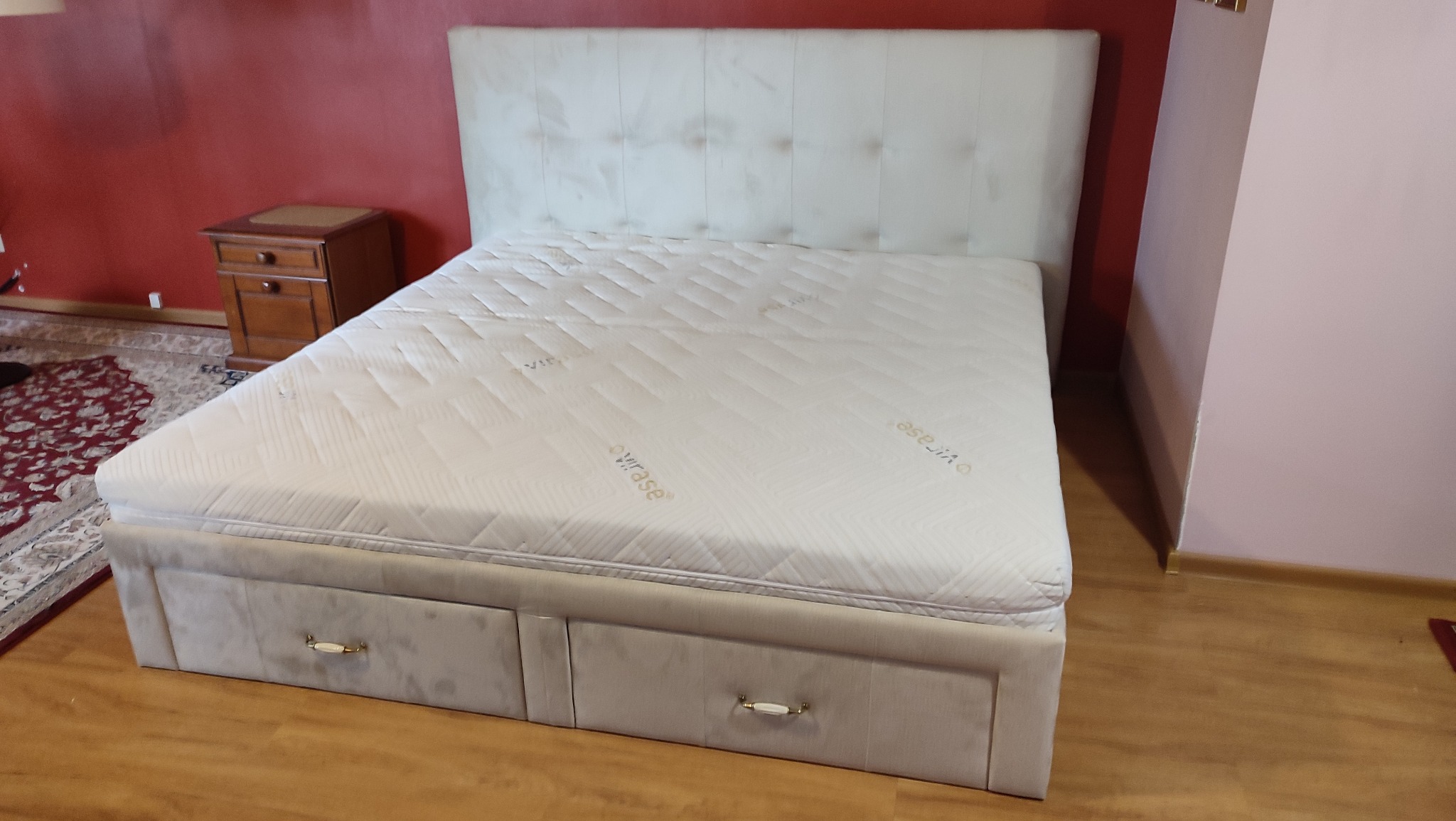 Beżowe łóżko tapicerowane z pojemnikiem i pikowanym zagłówkiem, widoczne na tle ściany w odcieniach czerwieni i różu. Obok szafka nocna i fragment dywanu.