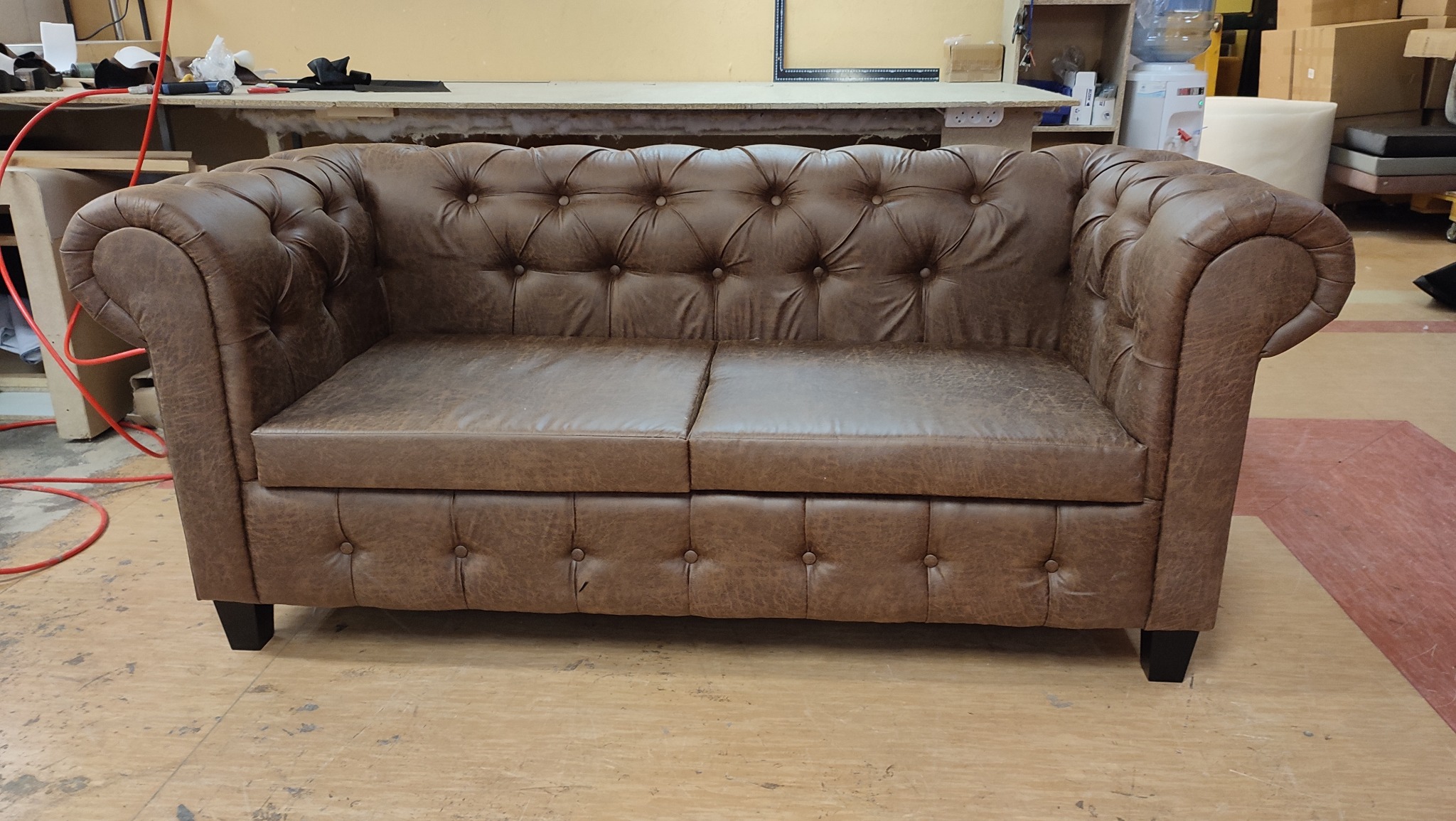 Brązowa, skórzana sofa typu chesterfield z głębokimi pikowaniami na oparciu i podłokietnikach, stojąca na drewnianej podłodze w warsztacie tapicerskim.