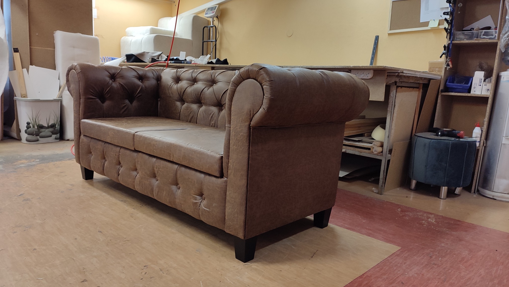 Brązowa sofa Chesterfield z guzikami, ustawiona w warsztacie tapicerskim. Widoczne narzędzia i materiały w tle. Charakterystyczny styl i wykończenie.