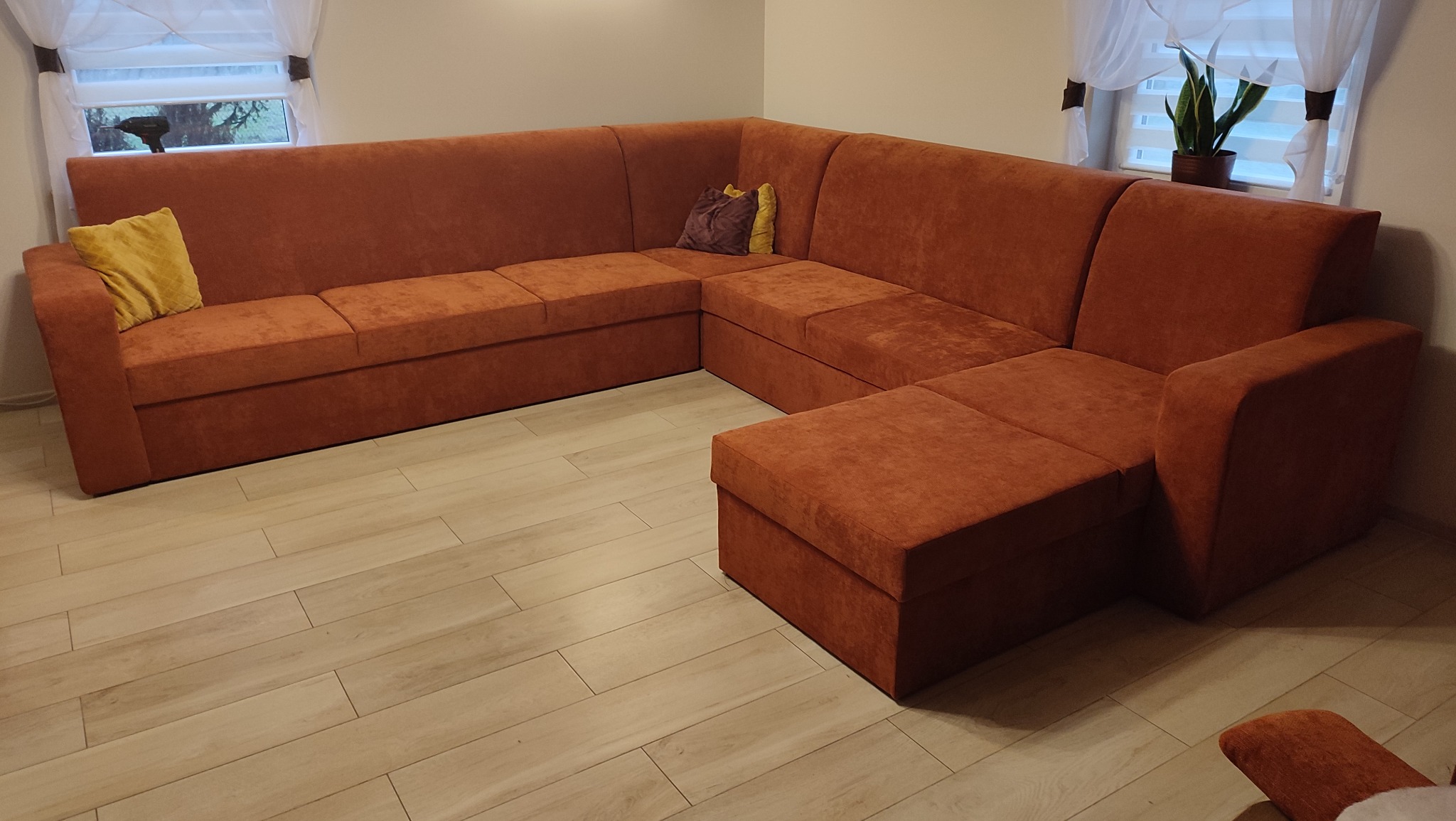 Pomarańczowy narożnik tapicerowany z poduszkami w salonie, widok z niskiego kąta, jasne panele podłogowe, okna z białymi firankami w tle.