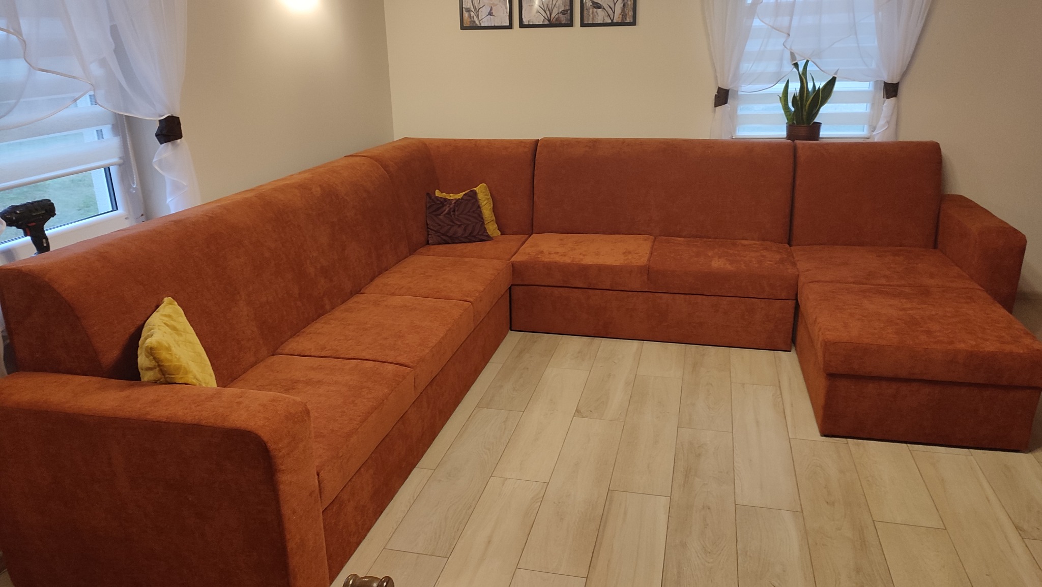 Pomarańczowy narożnik z funkcją spania, z poduszkami w salonie. W tle okno z firankami i roletami, na parapecie roślina doniczkowa. Na oparciu wiertarka.