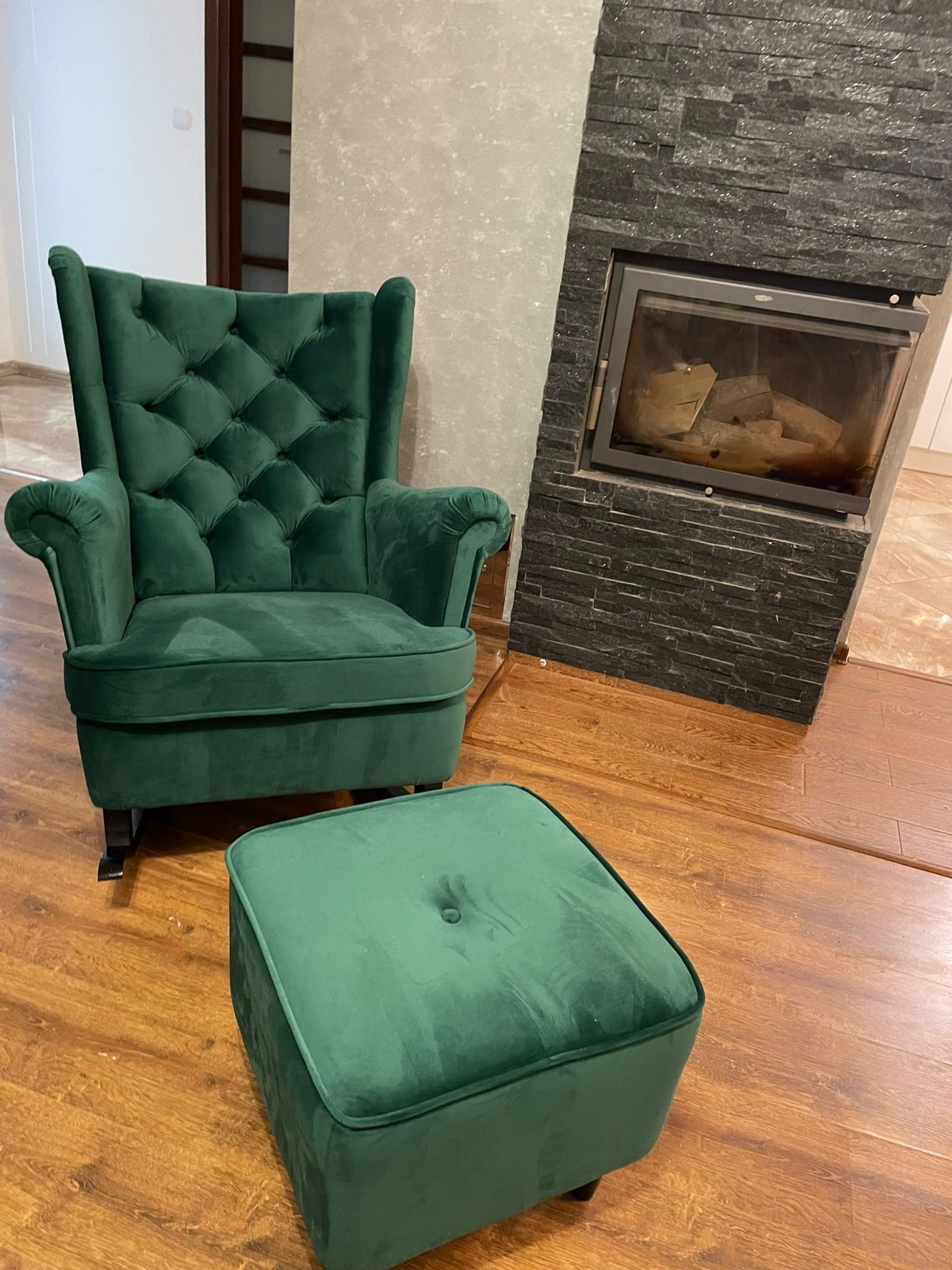 Szmaragdowy fotel uszak z podnóżkiem obitym welurem, na drewnianej podłodze, obok kominka z czarnej cegły. Elegancki design wnętrza.