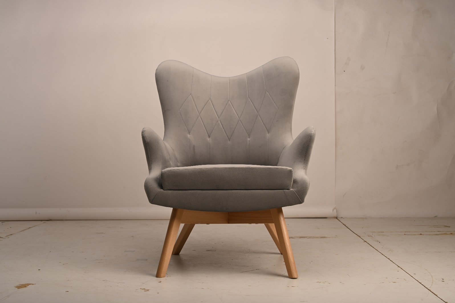 Szary, tapicerowany fotel z drewnianymi nogami na jasnym tle. Fotel ma charakterystyczne przeszycia w romby na oparciu i podłokietniki.