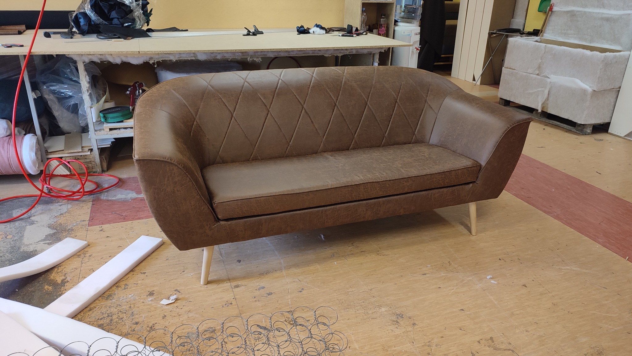 Gotowa, brązowa sofa z pikowaniem w romby, na jasnych nóżkach, w warsztacie tapicerskim. Widoczne sprężyny i pianka tapicerska.