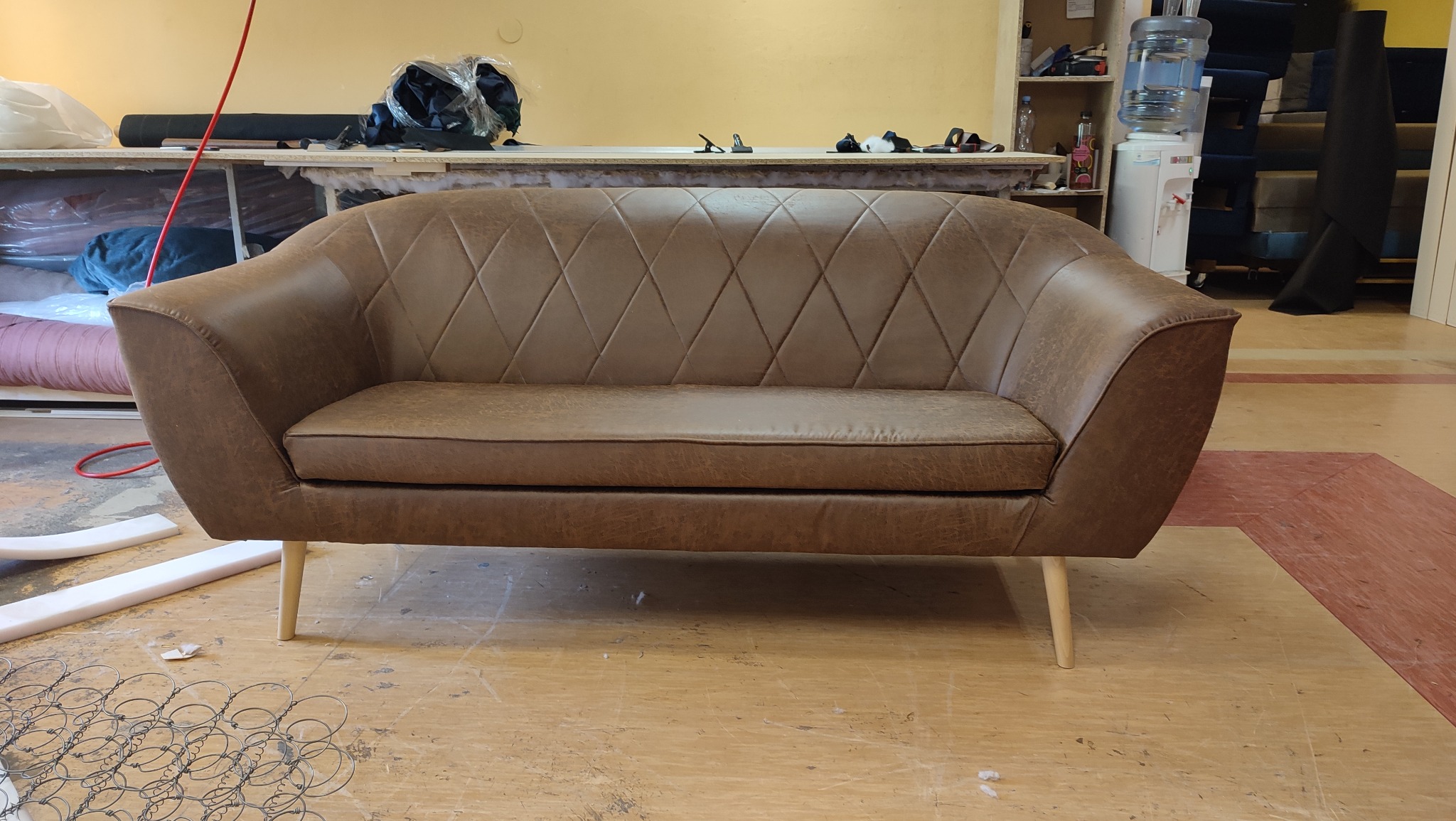 Brązowa sofa z pikowaniem w romby, na drewnianych nóżkach, w warsztacie tapicerskim. W tle materiały i narzędzia tapicerskie.