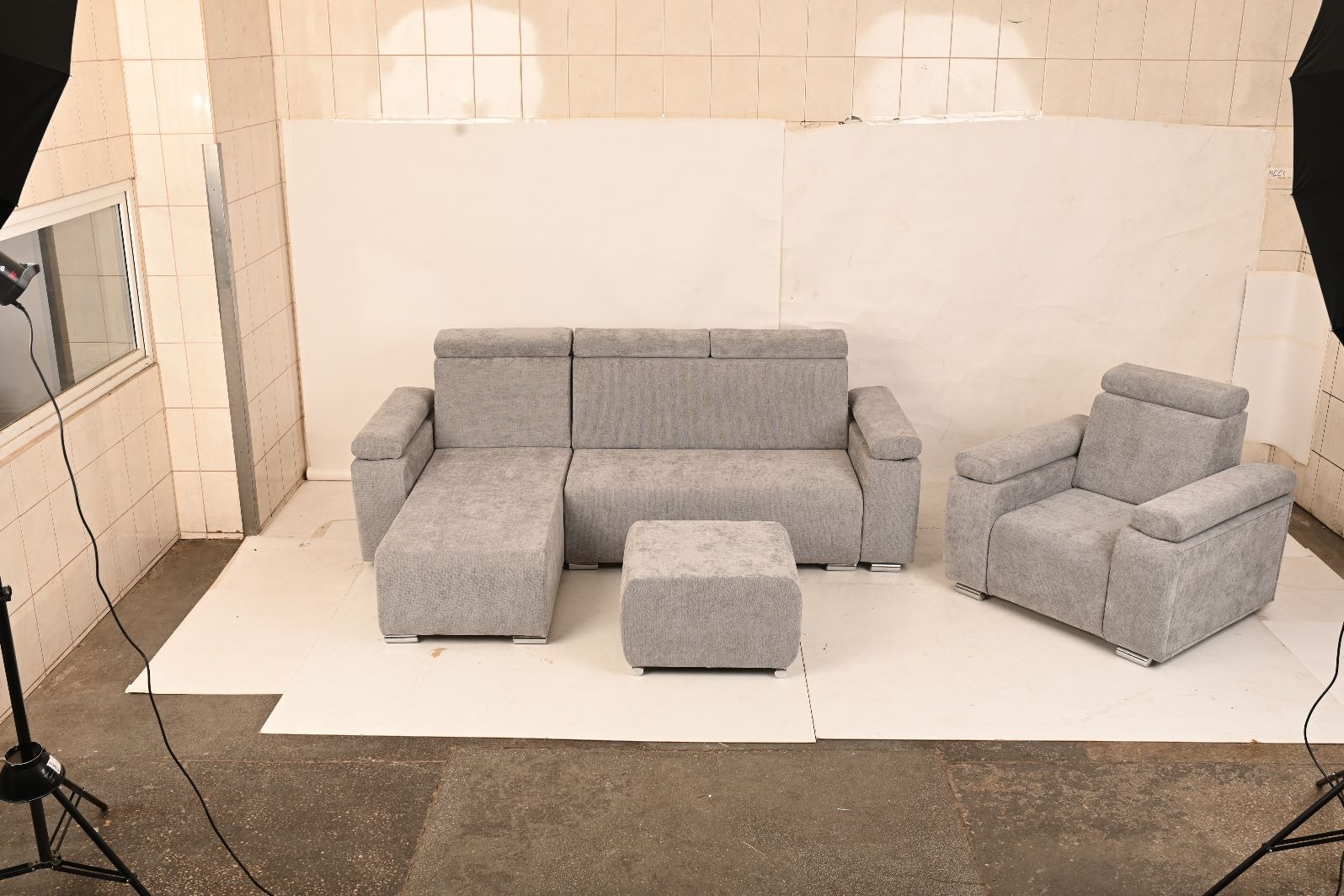 Komplet wypoczynkowy: szara sofa z szezlongiem, pufa i fotel. Nowoczesny design, minimalistyczne nóżki. Ustawiony w studiu fotograficznym z lampami.