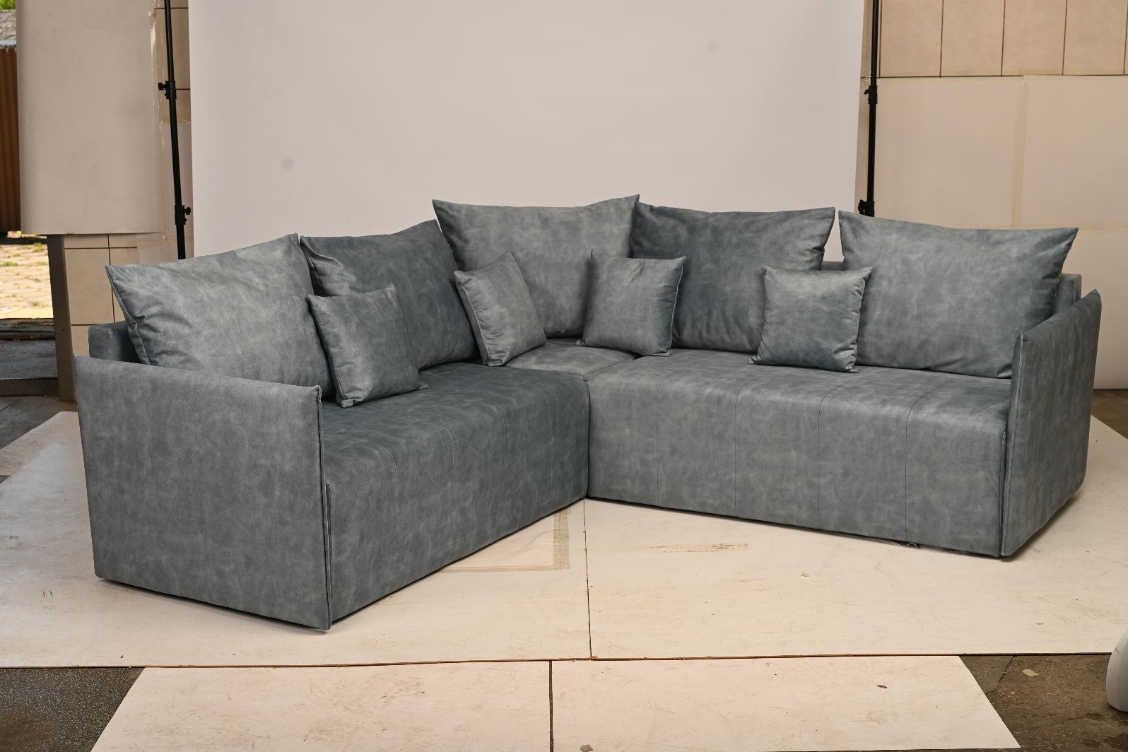 Szary, narożny zestaw wypoczynkowy z poduszkami, wykonany z materiału imitującego skórę, na jasnej podłodze. Minimalistyczny design, neutralne tło.