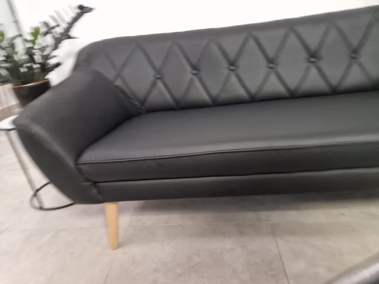 Czarna, skórzana sofa z pikowanym oparciem w romby i drewnianymi nóżkami. Obok stolik z rośliną. Ujęcie z dołu, podkreślające nowoczesny design mebla.