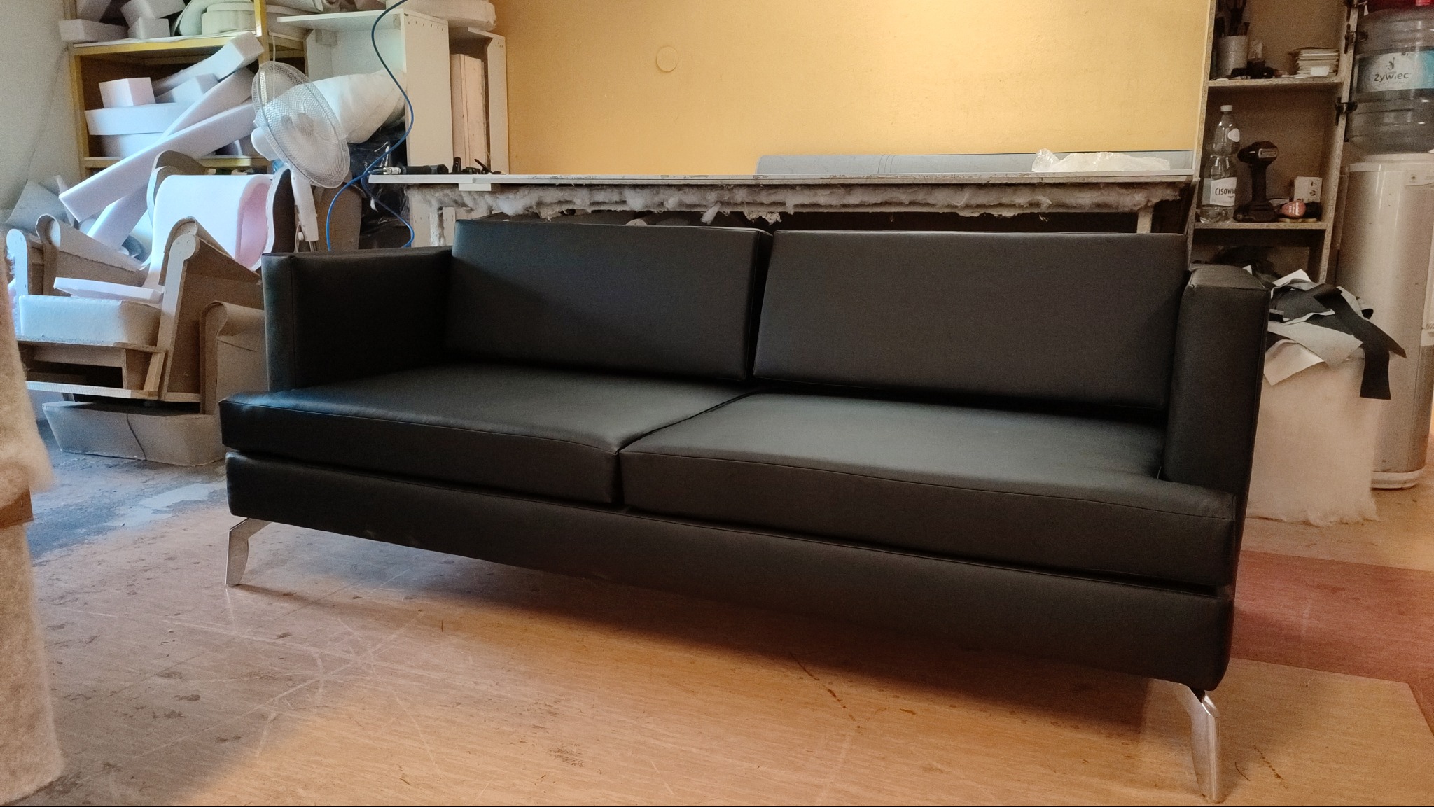 Nowoczesna, czarna sofa z ekoskóry na metalowych nóżkach, sfotografowana w warsztacie tapicerskim z widocznymi materiałami i narzędziami w tle.