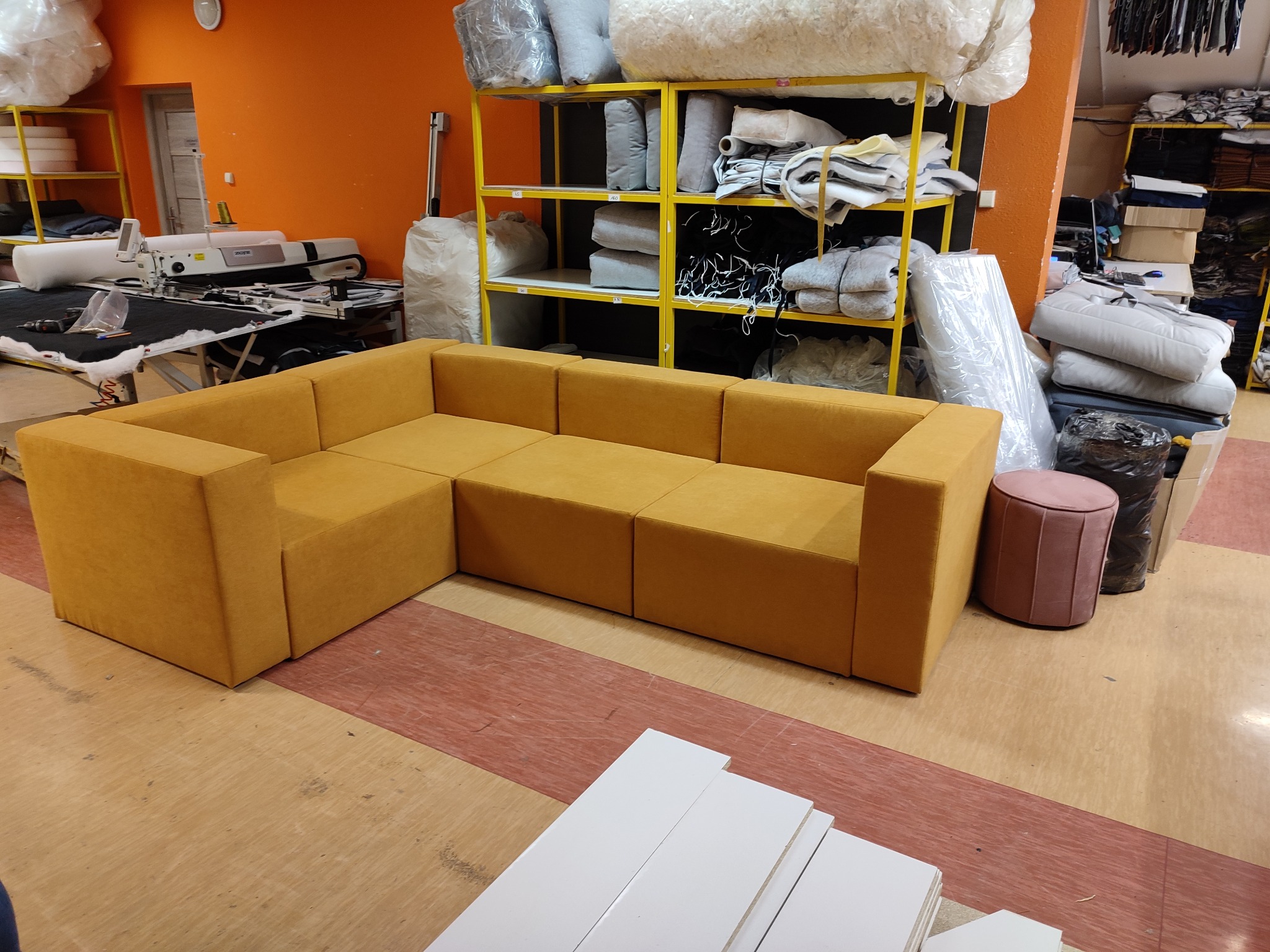 Kanciasta, modułowa sofa narożna w odcieniach musztardowej żółci, ustawiona w warsztacie tapicerskim z widoczną maszyną do szycia i regałami z materiałami.