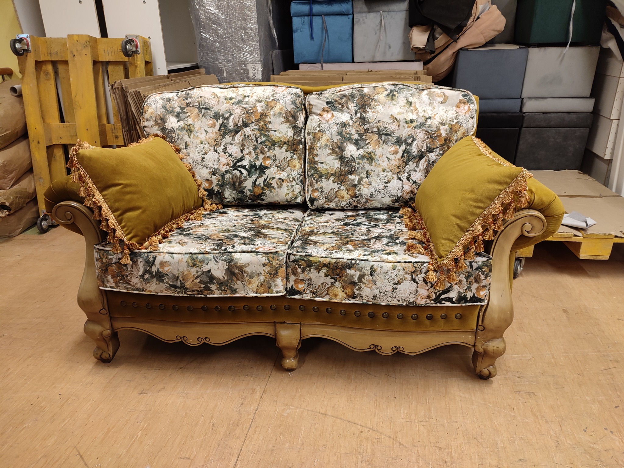 Dwuosobowa sofa w kwiaty z musztardowymi poduszkami i ozdobnymi frędzlami, ustawiona w magazynie na tle palet i kartonów.