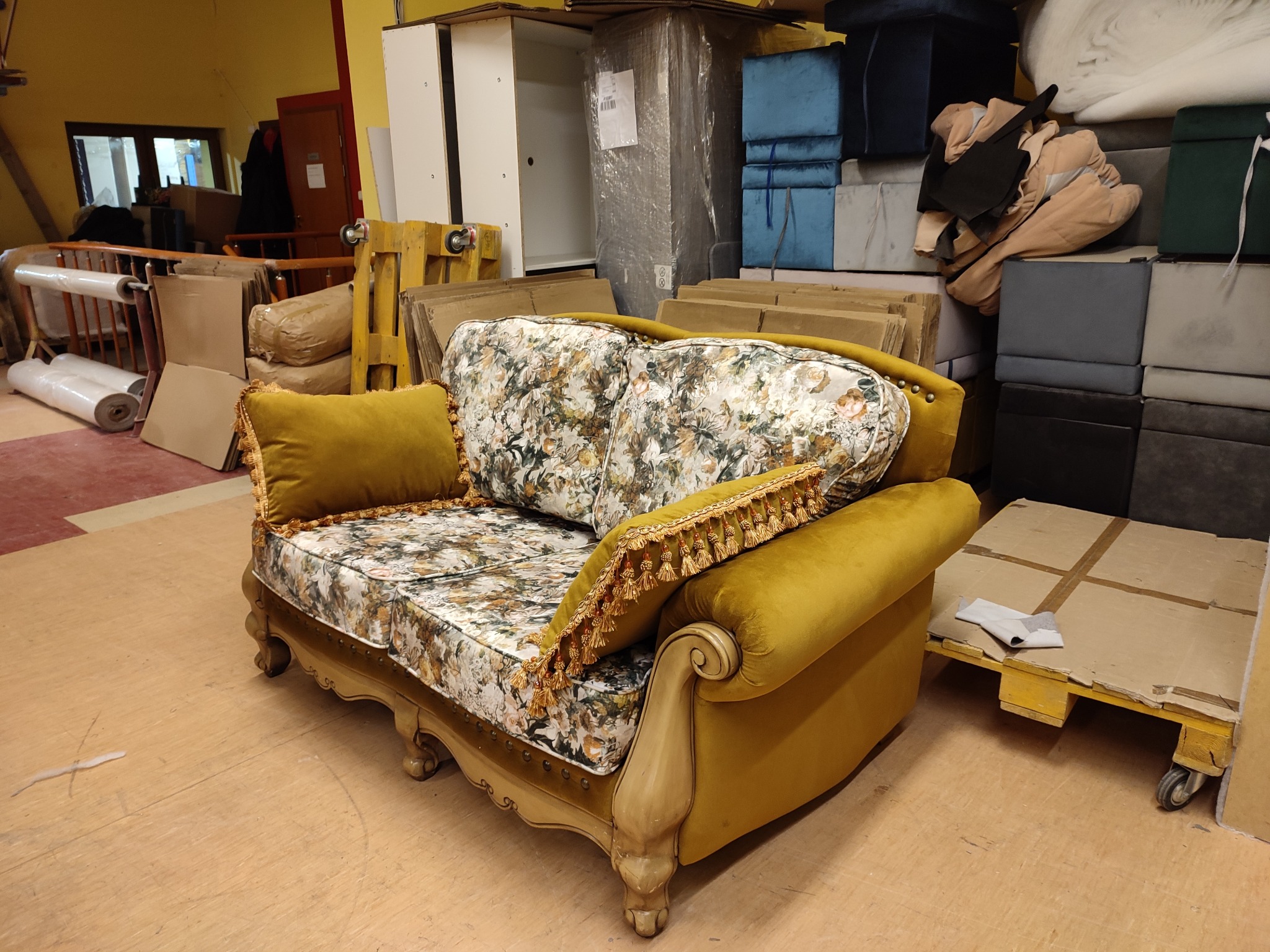Dwuosobowa sofa w stylu vintage z żółtymi, welurowymi bokami i poduszką, tapicerowana tkaniną w kwiaty, stoi w warsztacie tapicerskim na tle materiałów.
