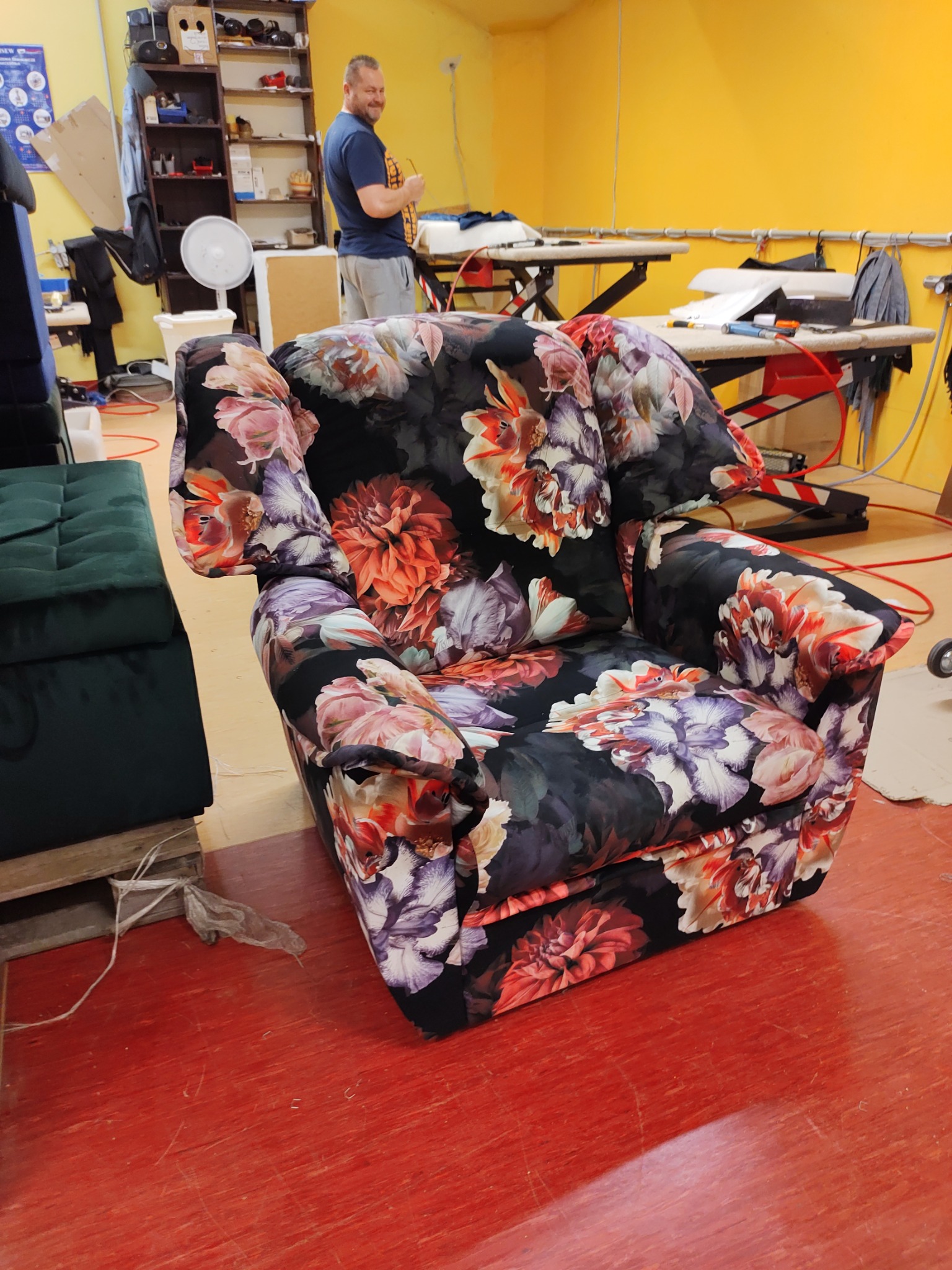 Ukończony fotel tapicerowany w kwiaty, stojący w warsztacie tapicerskim na czerwonej podłodze. W tle widoczny mężczyzna i narzędzia tapicerskie.
