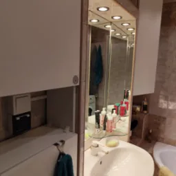 Łazienka z umywalką, szafką z lustrem i oświetleniem punktowym. W lustrze odbija się pralka i inne elementy wyposażenia łazienki. Widoczne płytki na ścianach.