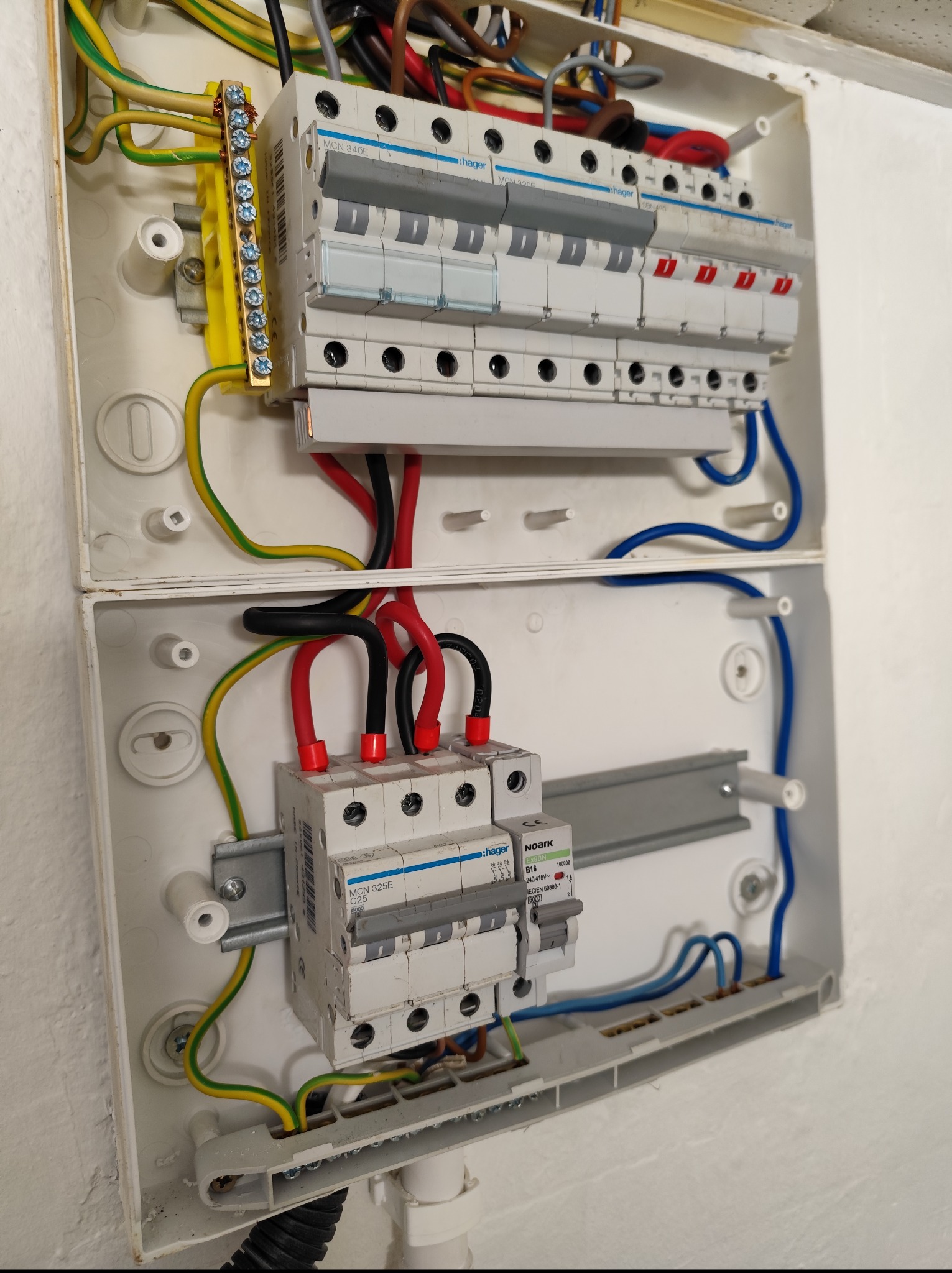 Otwarta skrzynka elektryczna z zainstalowanymi bezpiecznikami Hager i Noark, widoczne przewody w kolorach: żółto-zielonym, czarnym, czerwonym i niebieskim.