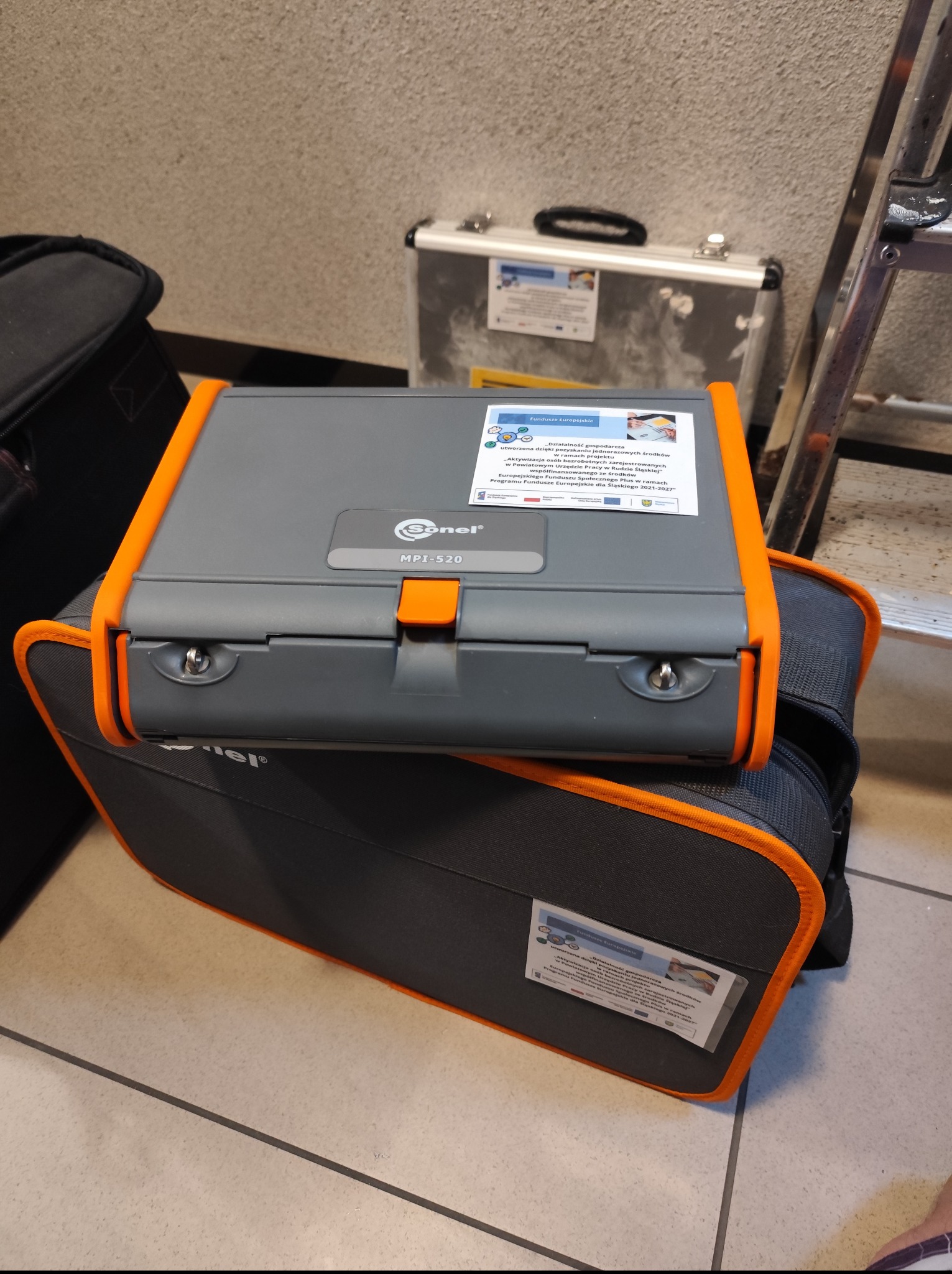Miernik Sonel MPI-520 na torbie narzędziowej, obok drabina i walizka. Urządzenie do pomiarów elektrycznych, na obudowie naklejka o dofinansowaniu z Funduszy Europejskich.